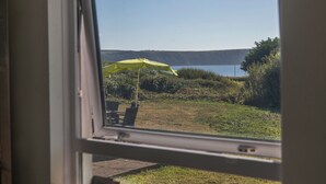 Vista a través de una ventana que muestra un área verde con una sombrilla amarilla, una silla y una masa de agua al fondo.