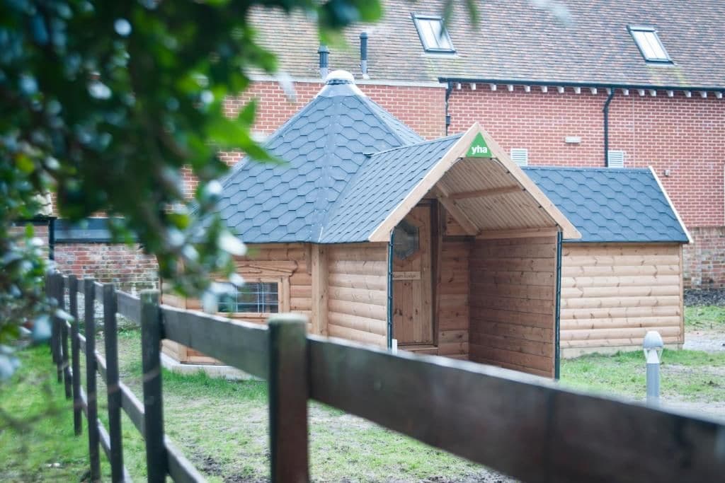 A wooden cabin with a sloped roof, a small porch, and a brick building in the background.