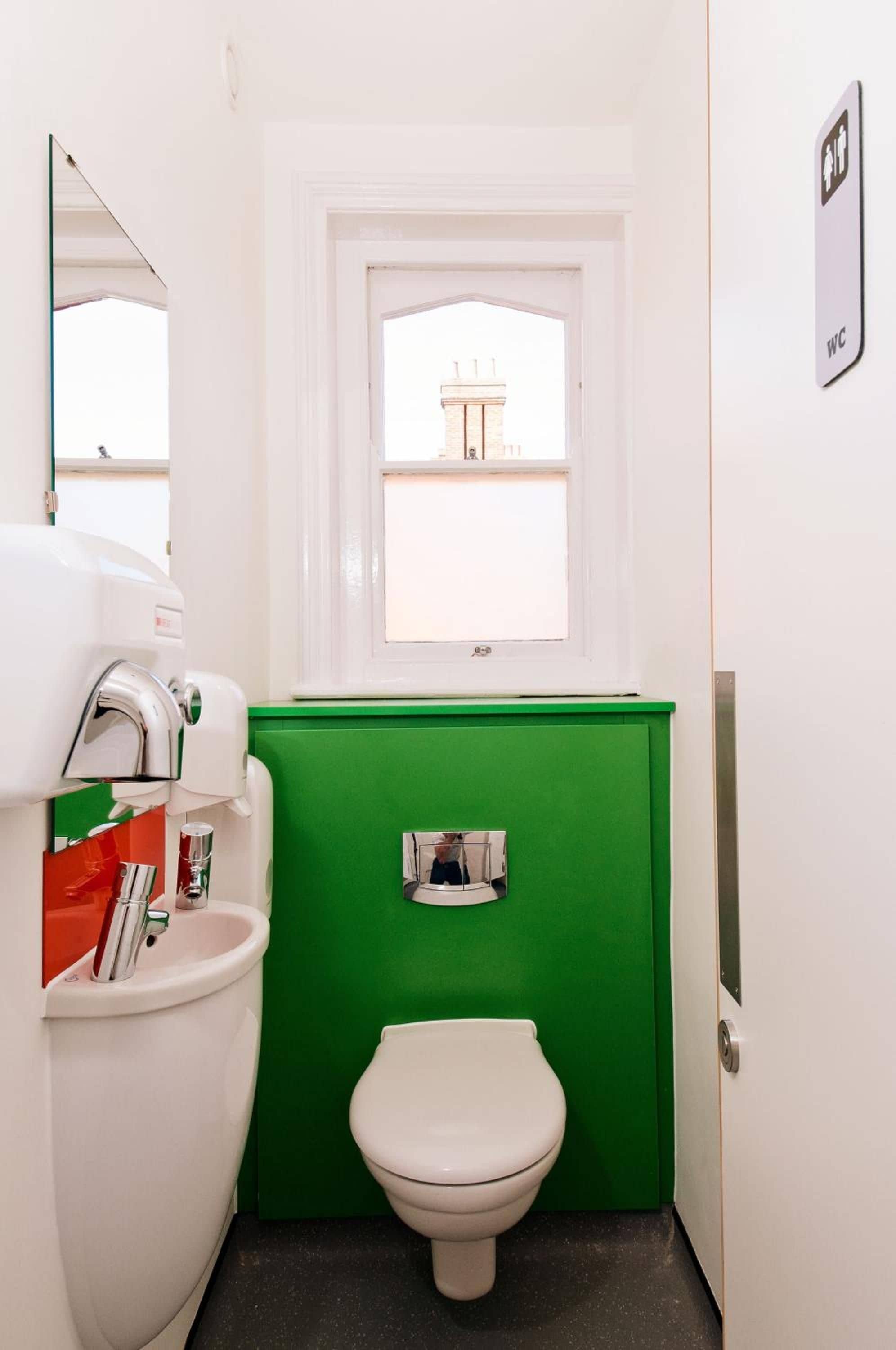 Un baño pequeño con inodoro blanco, separador verde, ventana y señalización de WC.