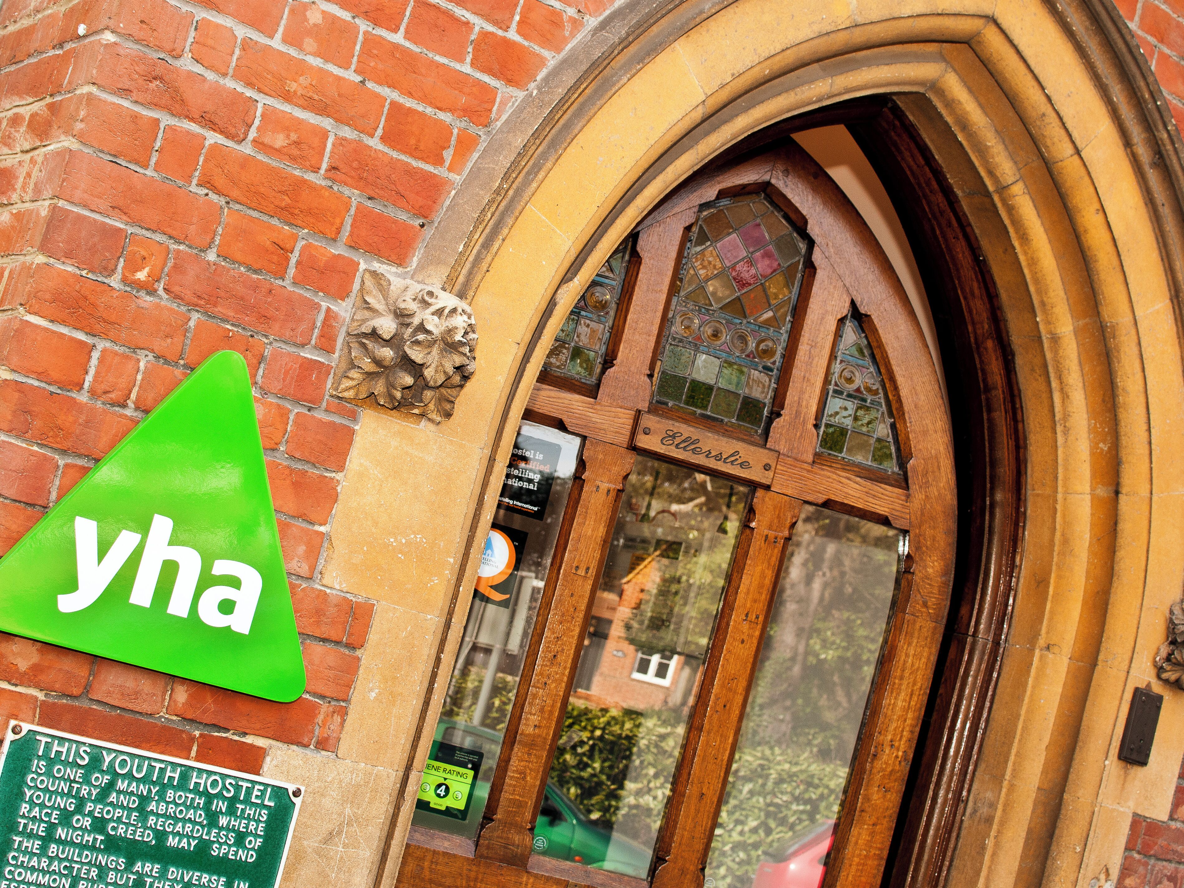 A hostel entrance with a green triangular sign, a wooden door with a glass window, and a brick wall.