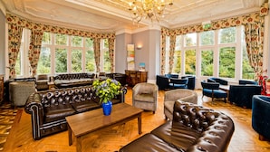 A spacious lounge with leather sofas, a wooden coffee table, and a chandelier. Large windows with floral curtains offer views of greenery.