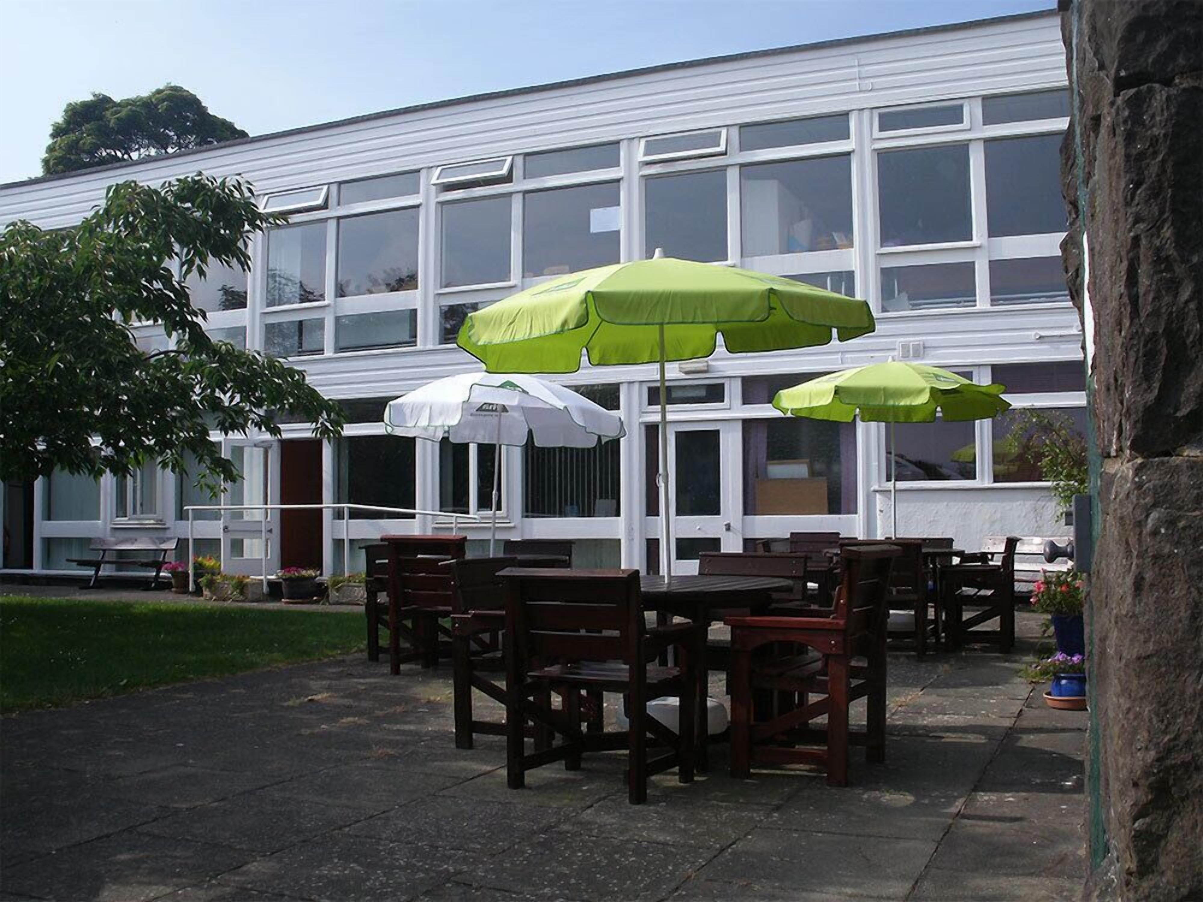 An outdoor patio area with wooden tables and chairs, covered by green and white umbrellas, in front of a building with large windows.