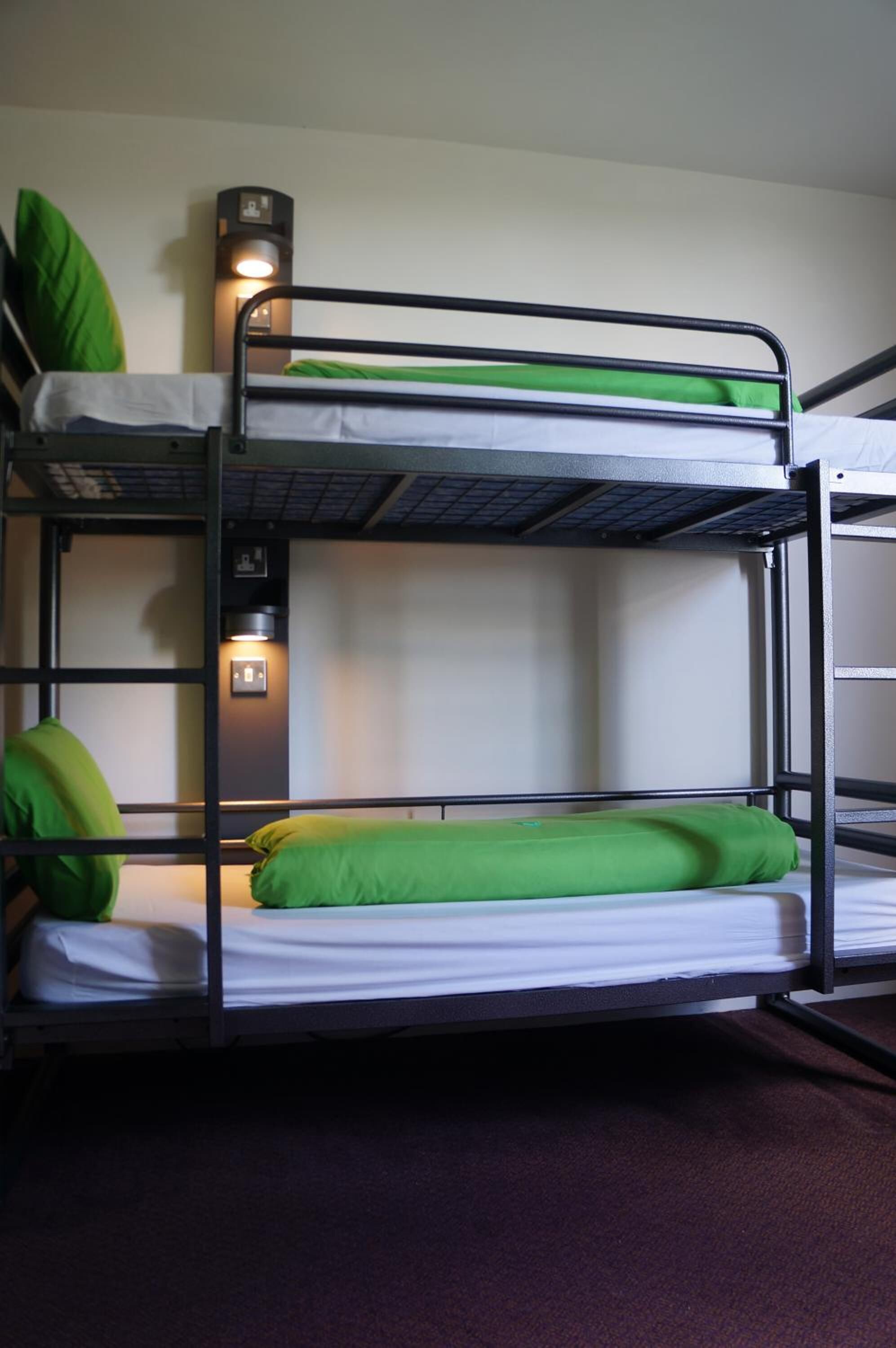 Dormitorio con literas, ropa de cama verde y una lámpara fijada a la pared.