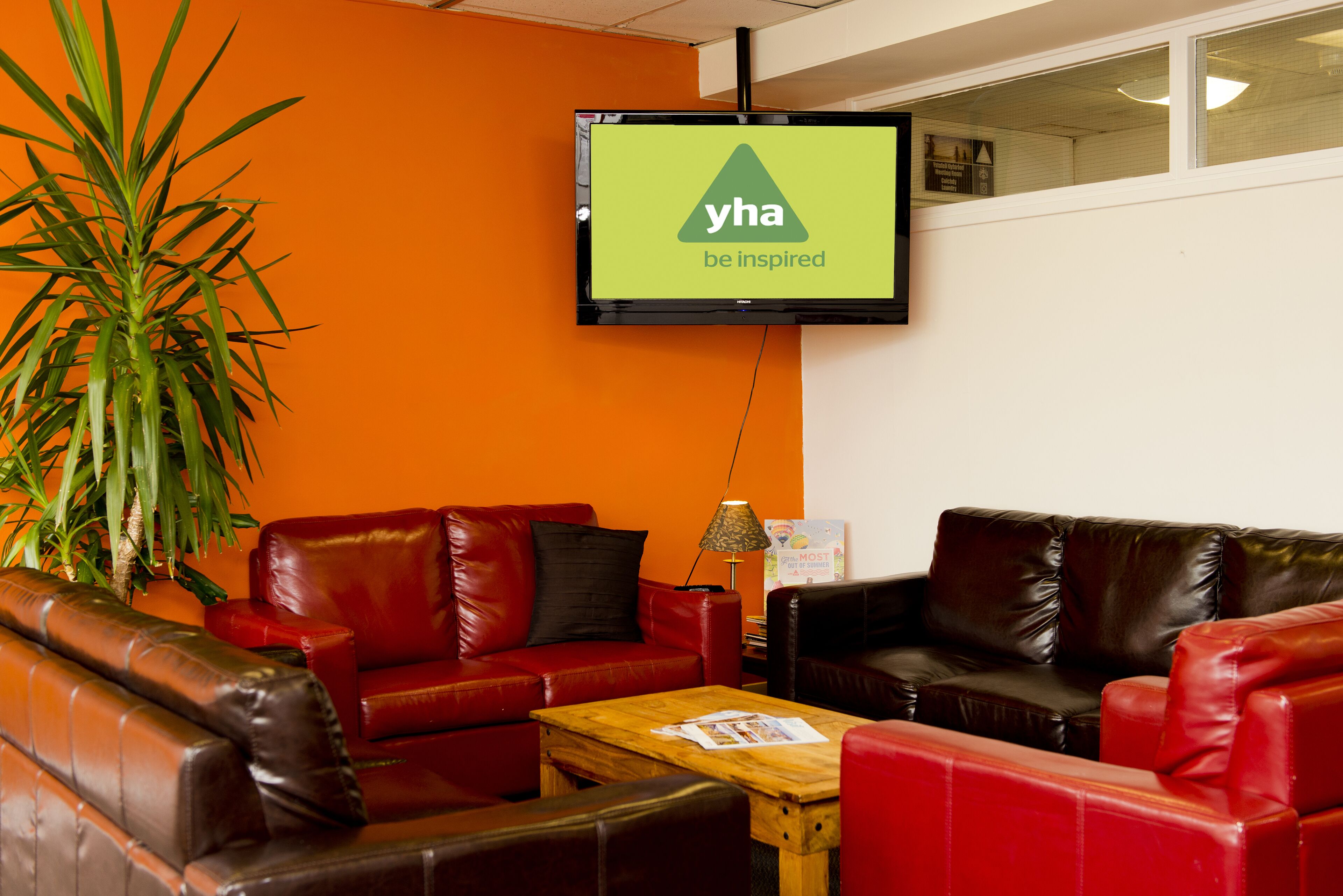 A room with red leather sofas, a wooden coffee table, and a large potted plant. A television screen displays the logo "yha be inspired".