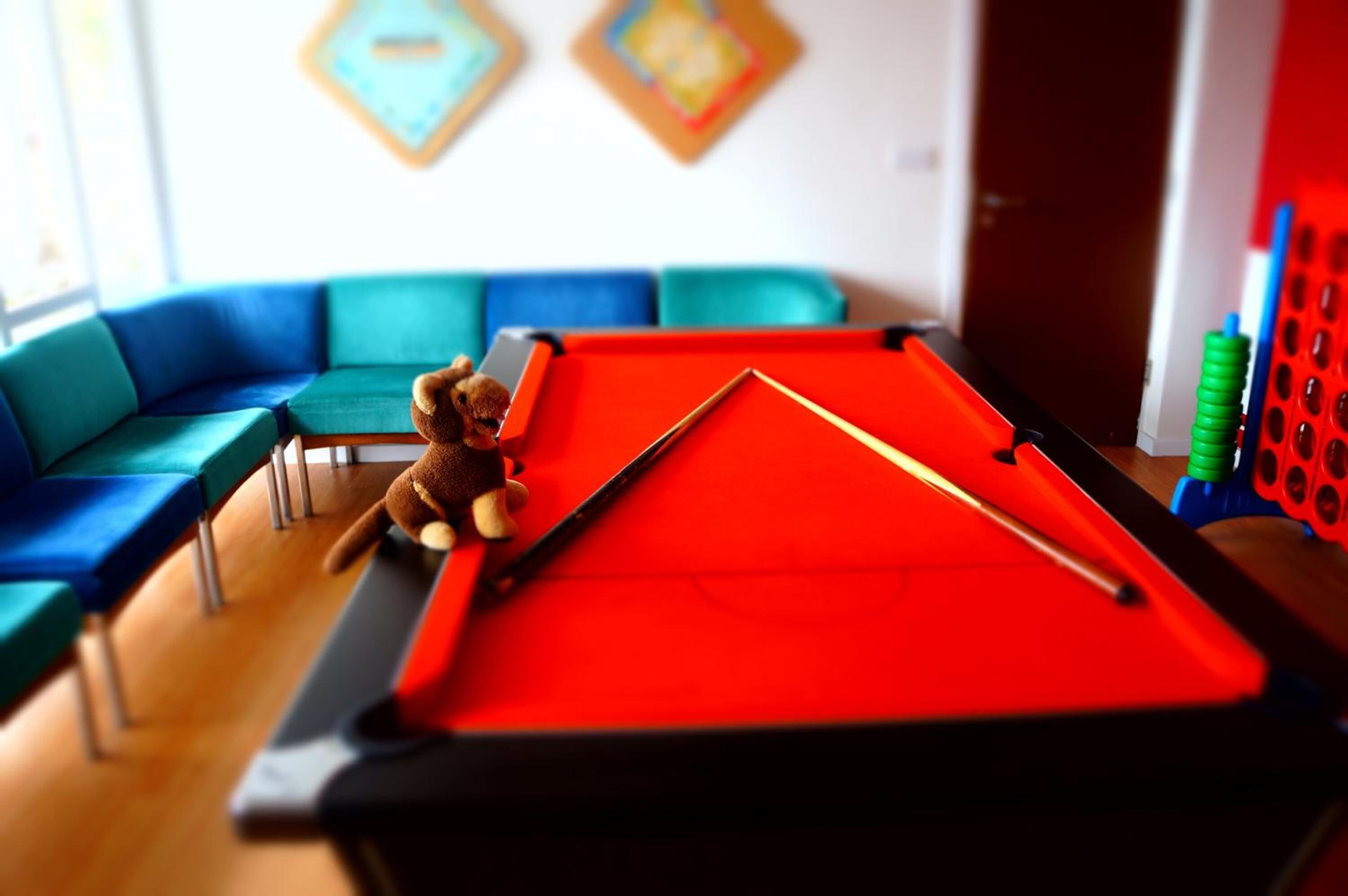 A pool table with a red felt surface, a brown teddy bear, and a blue sofa in the background.