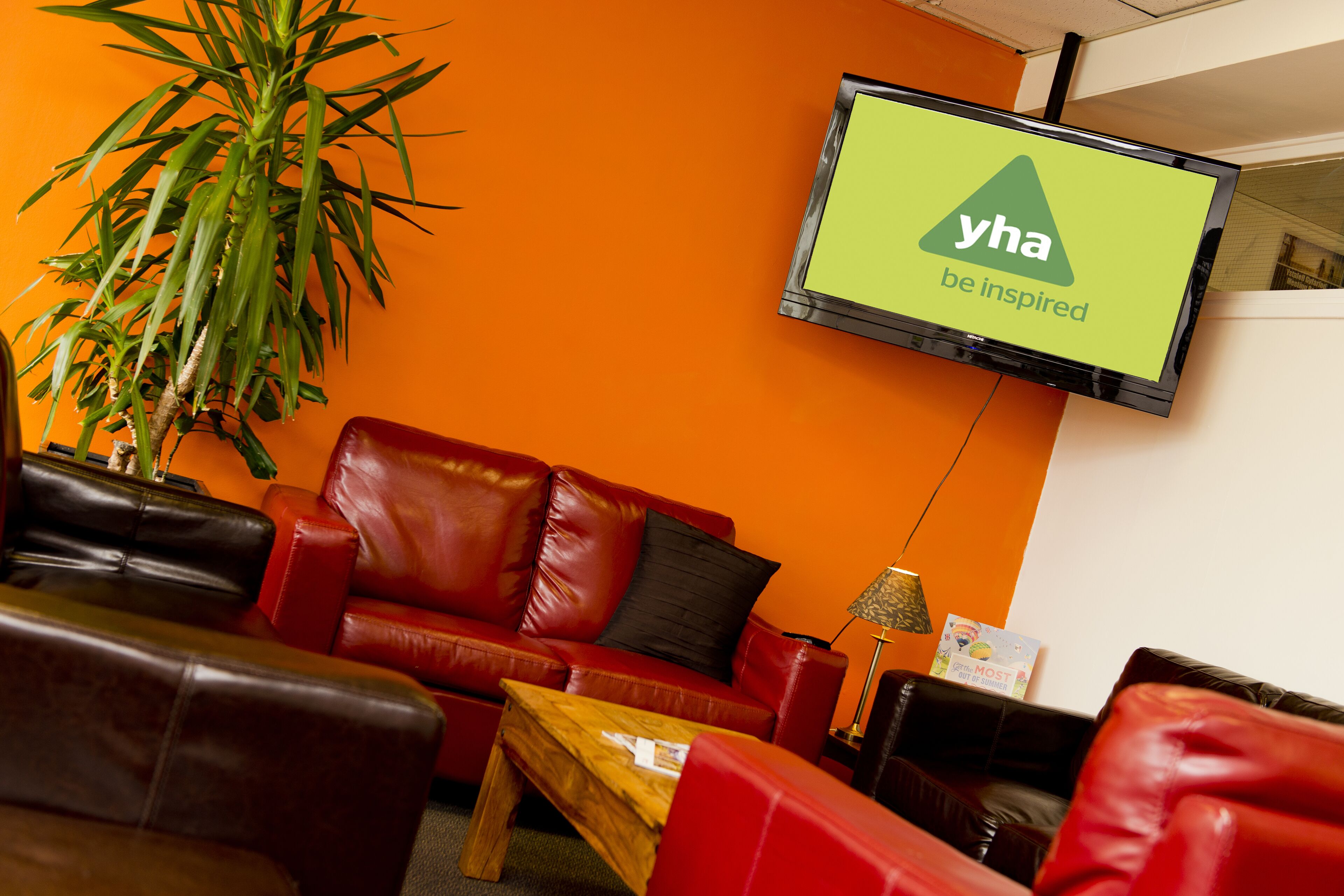 Un salon moderne avec un canapé en cuir rouge, une table basse en bois et une grande plante en pot. Un téléviseur à écran plat, fixé au mur, affiche le logo « yha be inspired ».