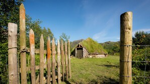 Una valla de madera rústica con una pequeña construcción de madera al fondo, rodeada de vegetación y colinas.
