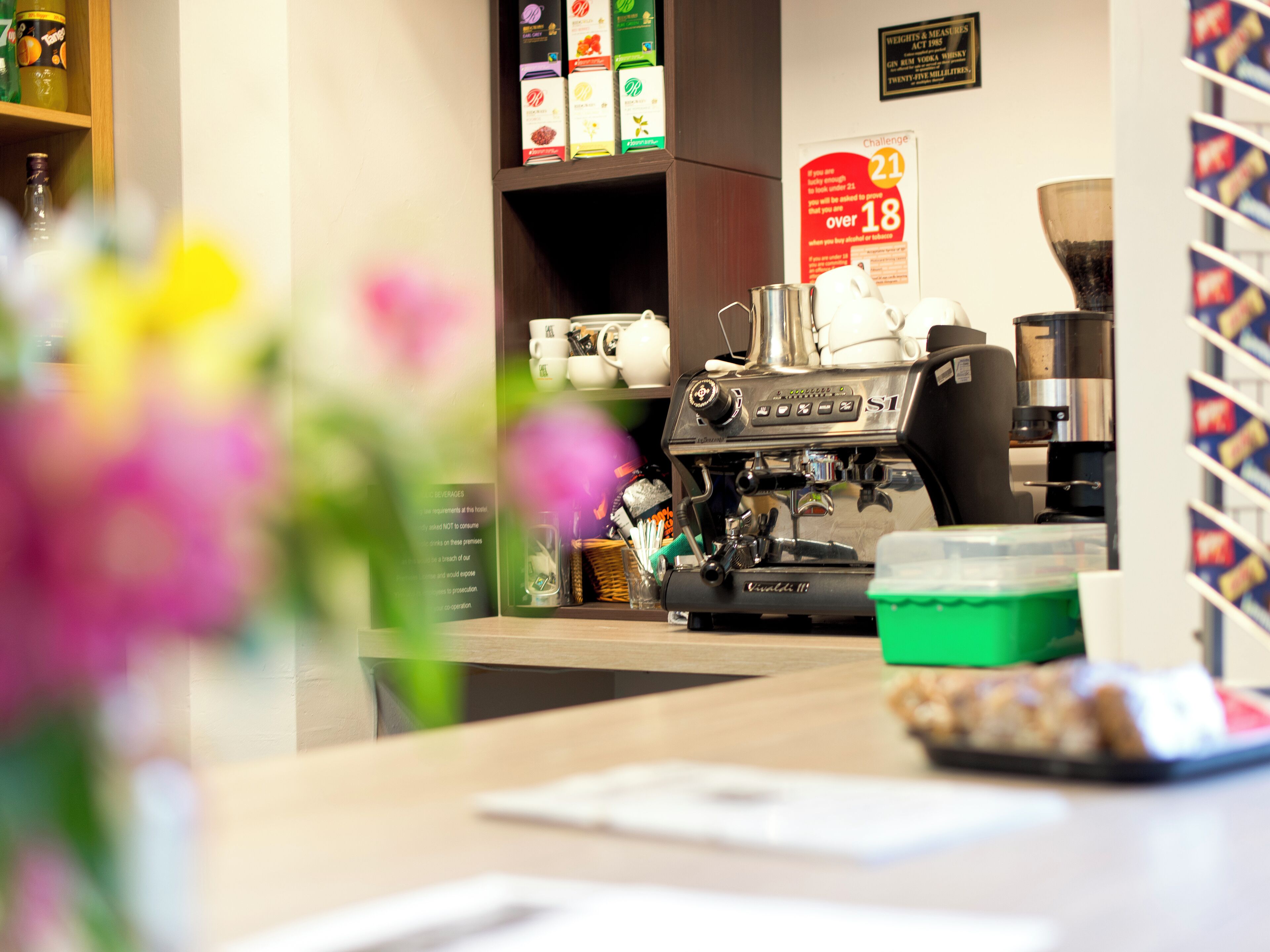 Un interior de cafetería con una máquina de espresso, una barra con un recipiente verde y estantes con diversos artículos.