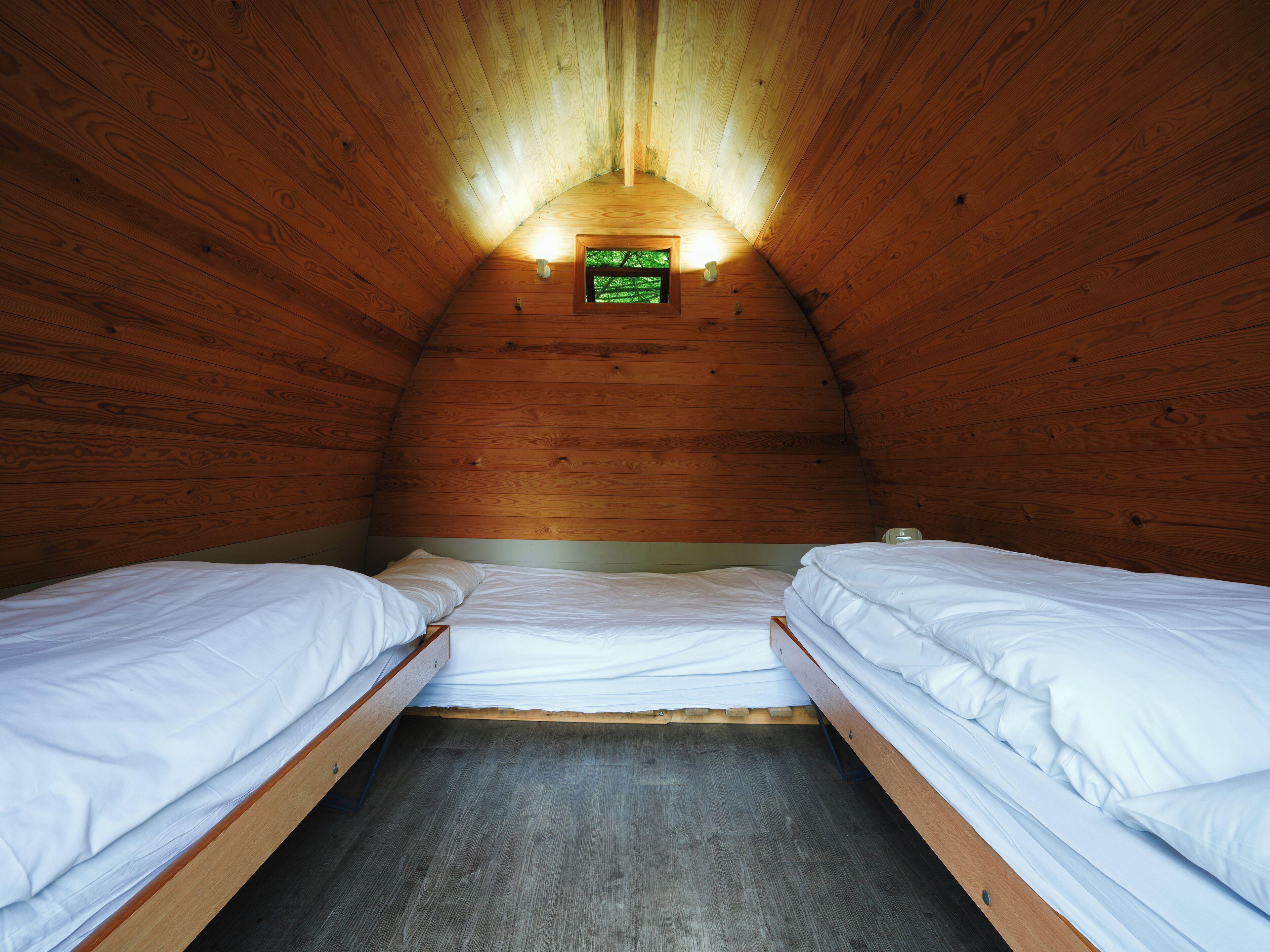 Una habitación pequeña y acogedora con paredes de madera y techo inclinado. Hay dos camas individuales con ropa blanca y una ventana pequeña al fondo de la habitación.