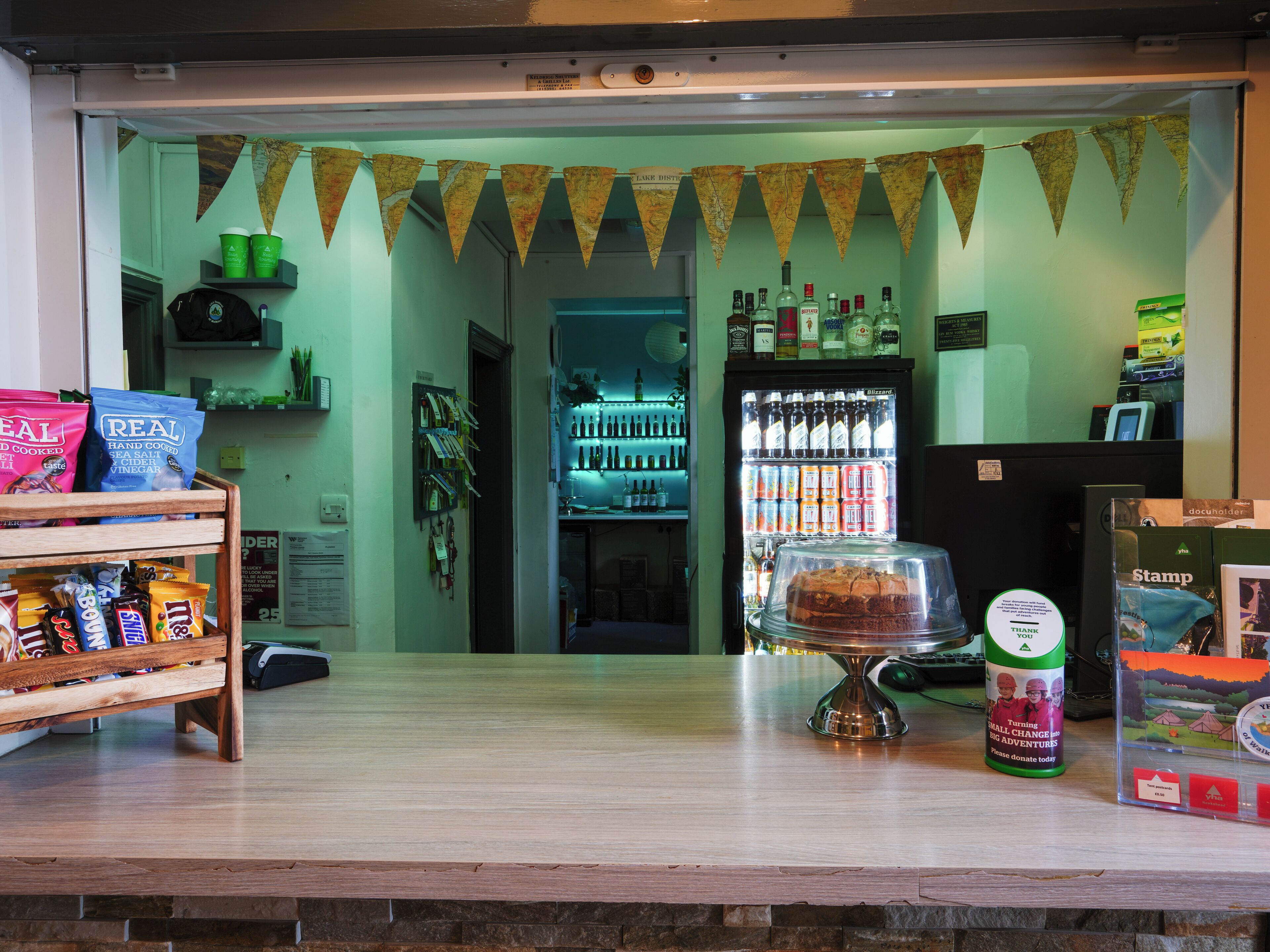 Una zona de bar con mostrador, estantes con aperitivos, un expositor refrigerado y un buzón de donaciones.