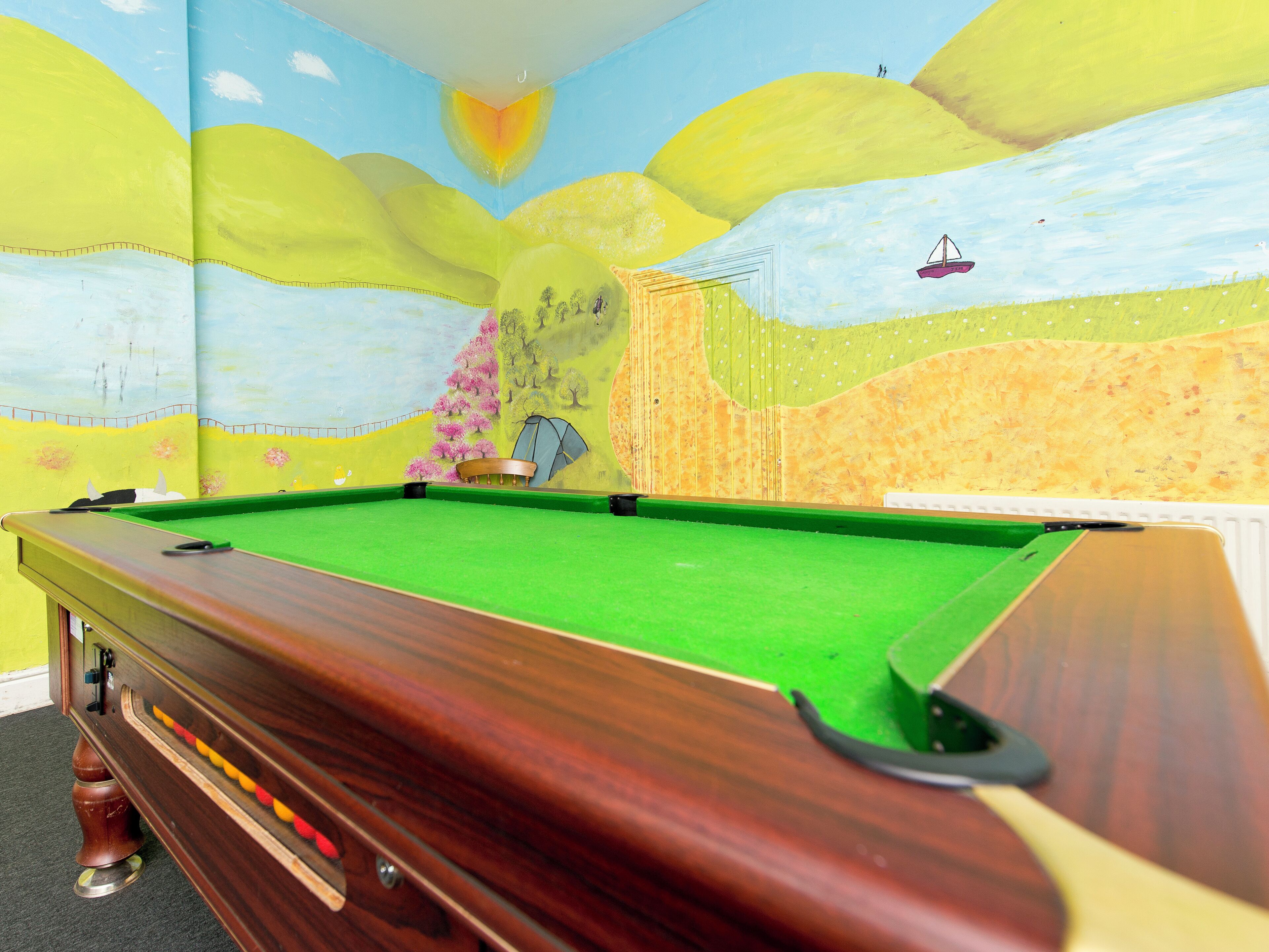 Een biljarttafel in een ruimte met een muurschildering van een landschap en een zeilboot op de muur.