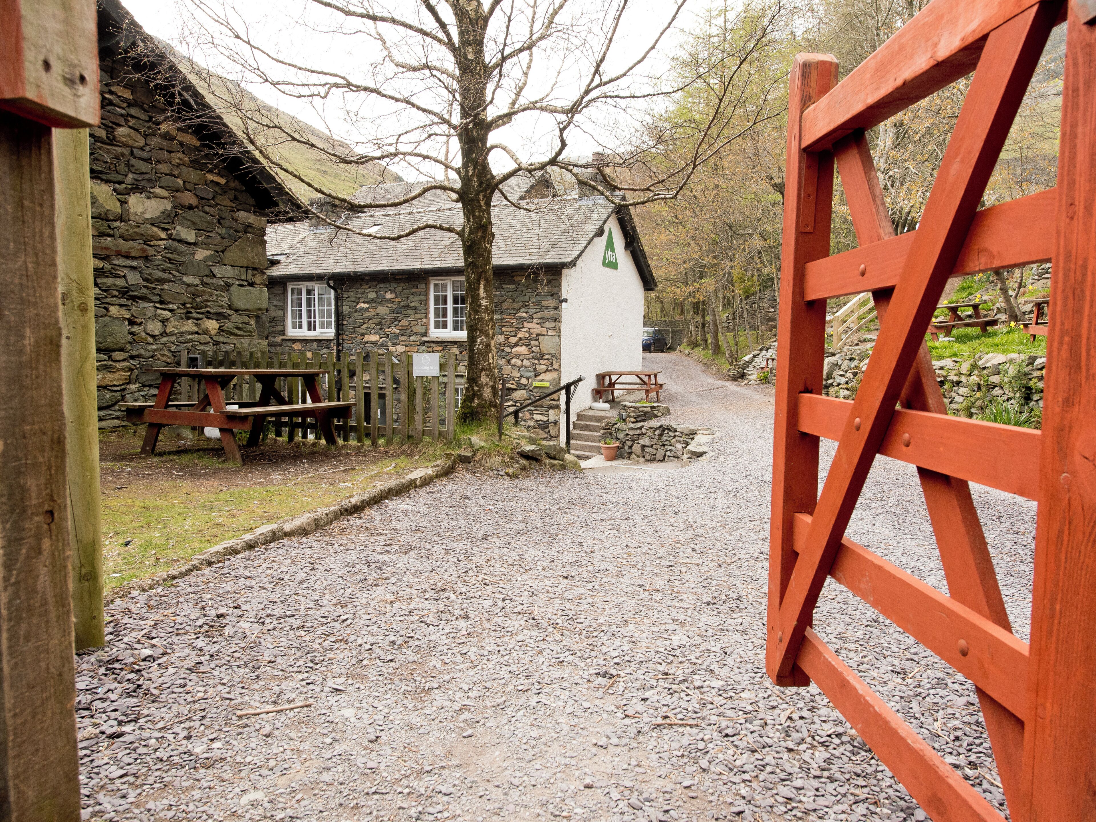 A rustic stone building with a white sign, a gravel path leading to a wooden gate, and picnic tables.