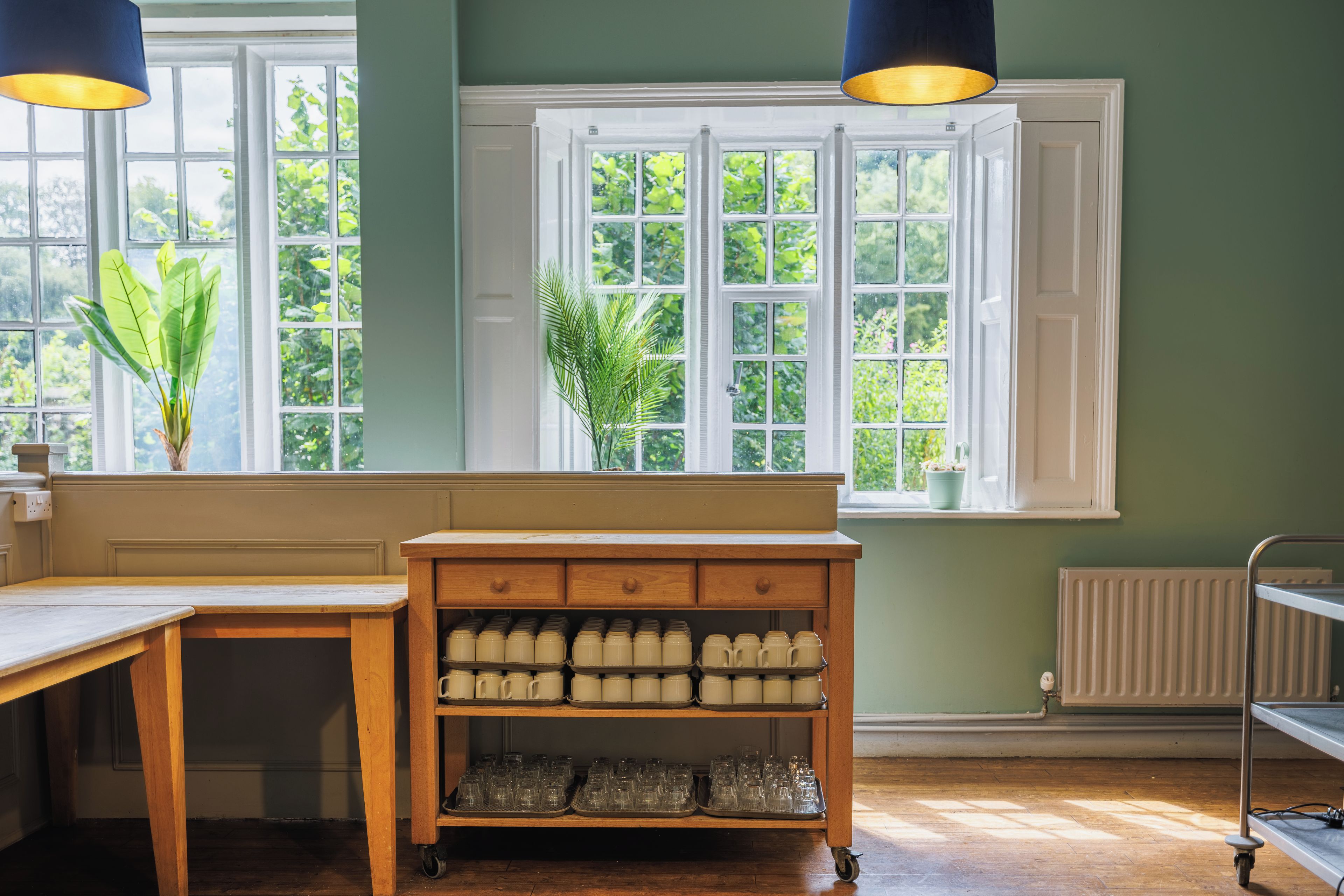A kitchen with a wooden bench, a rolling cart with mugs, and a window with a view of greenery.