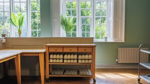 A kitchen with a wooden bench, a rolling cart with mugs, and a window with a view of greenery. - YHA Ilam Hall - Hostel (Ashbourne)