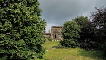 Un edificio in pietra con uno stile architettonico tradizionale, circondato da una vegetazione lussureggiante e un vialetto erboso che conduce alla costruzione.