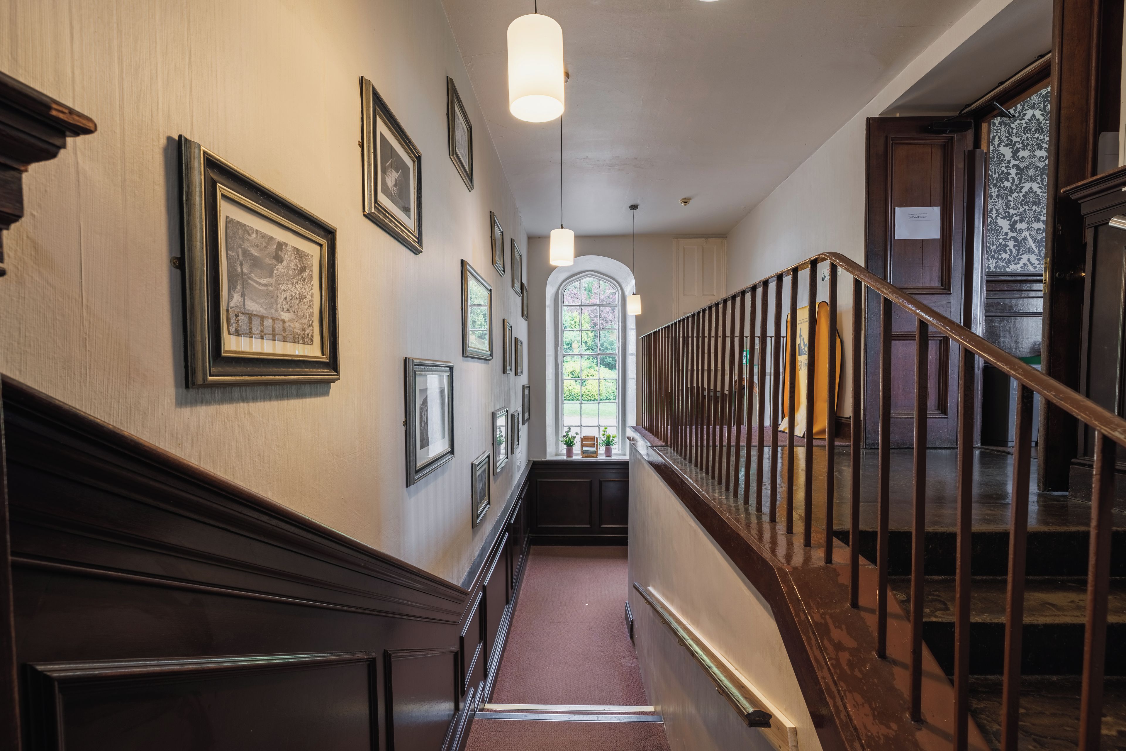 A hallway with framed pictures on the walls, a wooden staircase, and a window with a view of greenery.