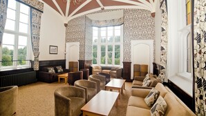 A spacious room with a high ceiling, patterned wallpaper, and large windows offering a view of the outdoors. There are multiple seating areas with sofas and armchairs, and a wooden table in the center. - YHA Ilam Hall - Hostel (Ashbourne)