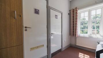 Una habitación con un armario de madera, una puerta con el letrero de baño, un radiador y una ventana con cortinas.
