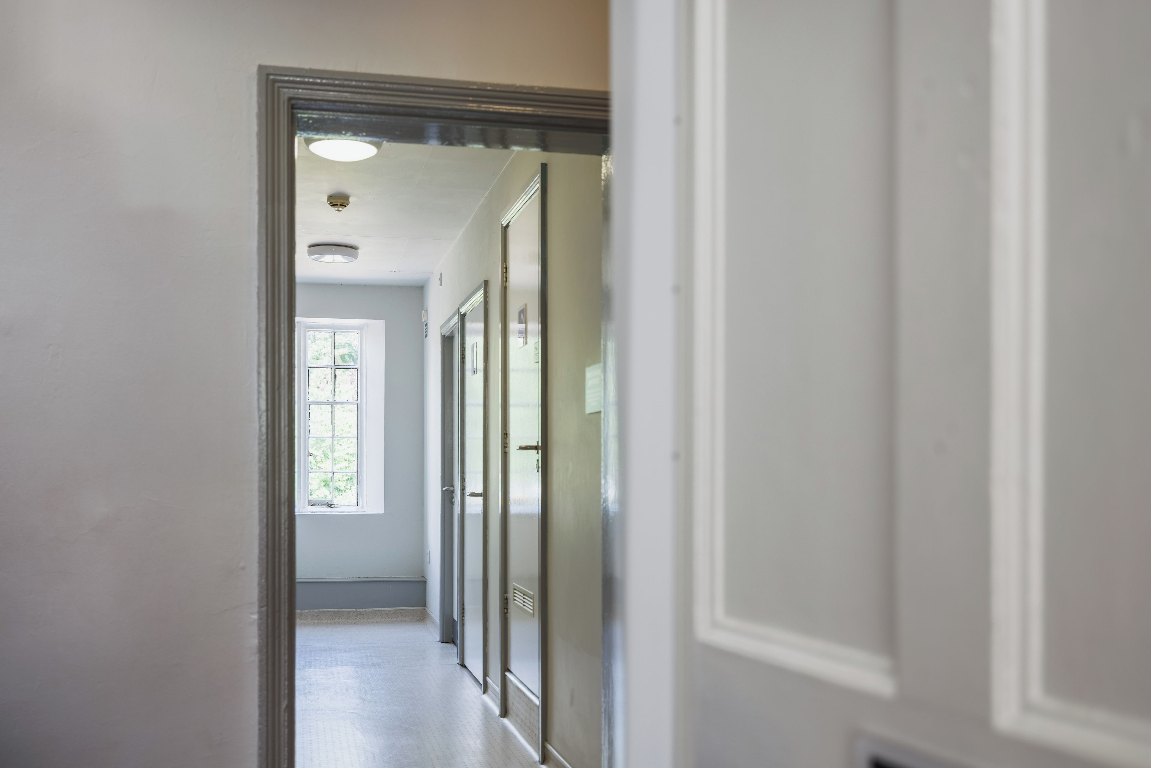 Corredor com porta que dá para um quarto, janela com vista para área verde e luminária no teto.