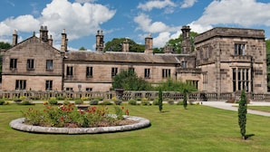 A historic stone building with multiple chimneys, a well-maintained lawn, and a circular flower bed. - YHA Ilam Hall - Hostel (Ashbourne)