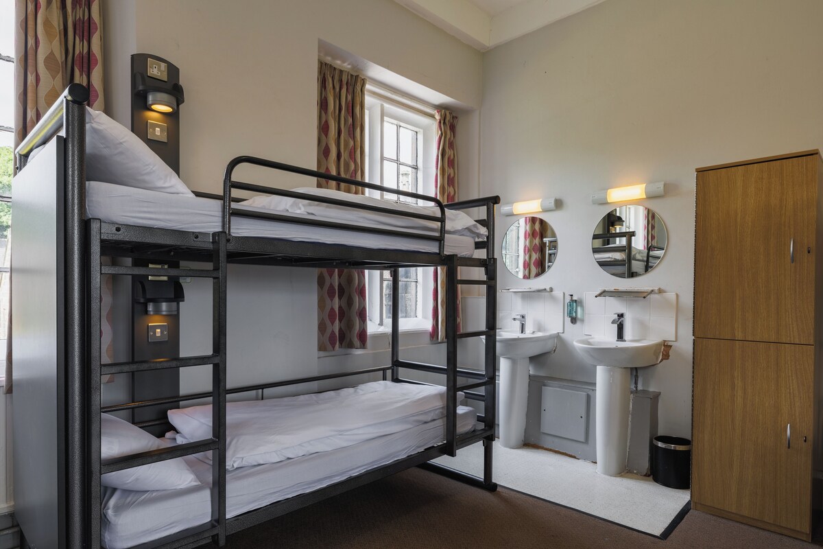 A bunk bed room with a sink and mirror, a wooden wardrobe, and a window with curtains.
