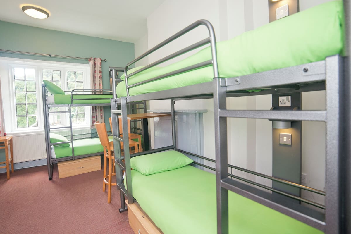 A bunk bed room with green bedding, a wooden chair, and a window with a view.
