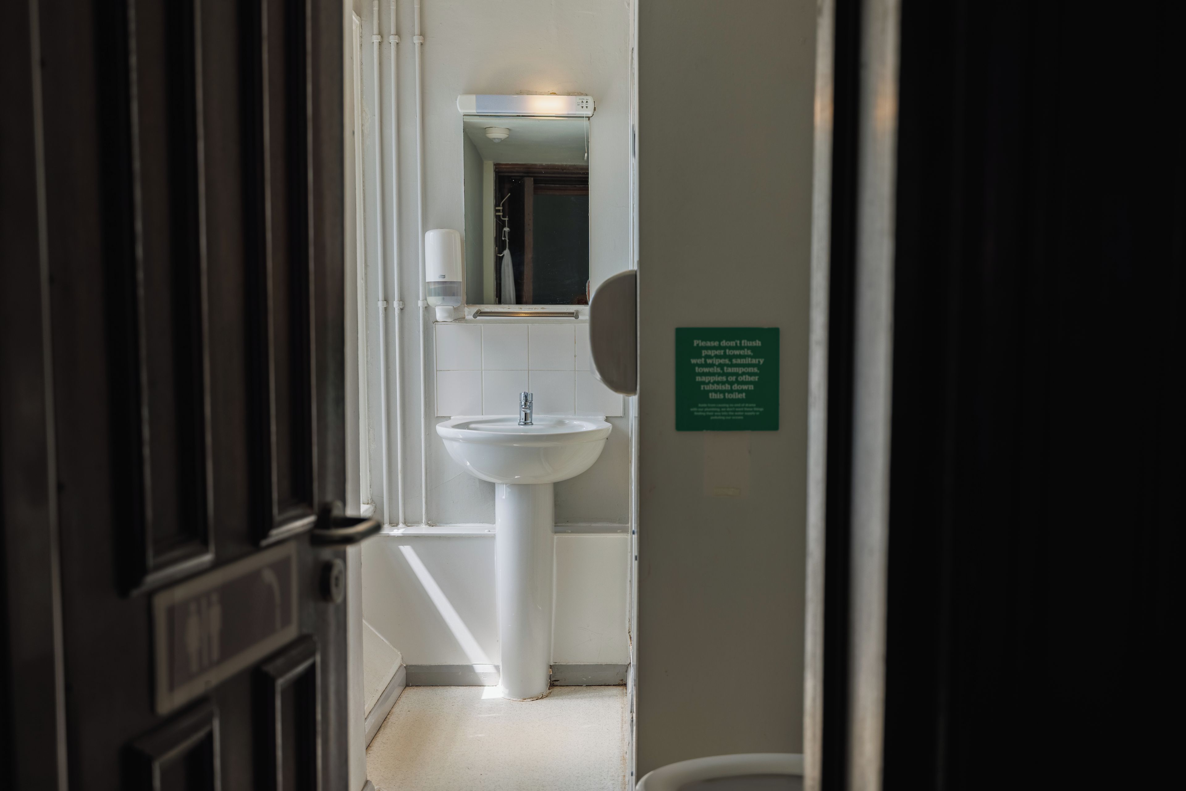A narrow hallway leading to a bathroom with a sink and a hand soap dispenser. A sign on the wall requests not to flush paper towels.