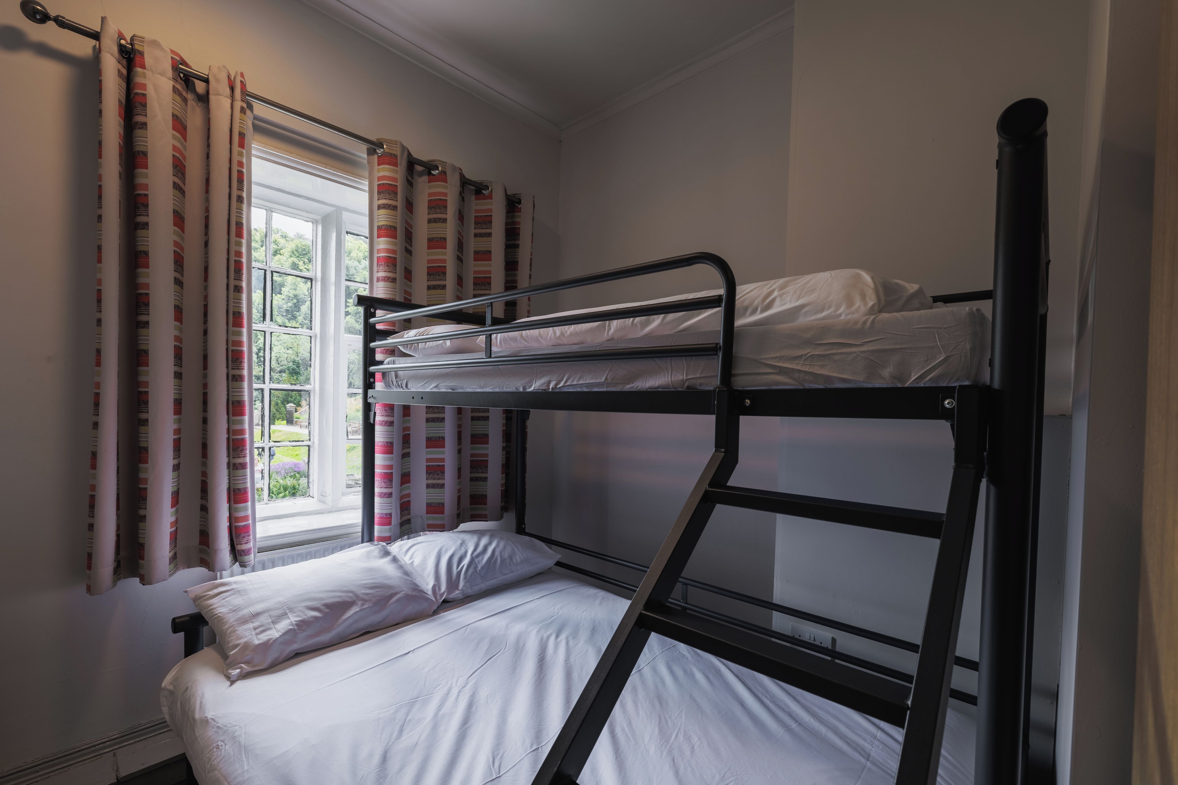 Ein Stockbett mit weißer Bettwäsche, ein Fenster mit Vorhängen und Blick auf Grünfläche draußen.