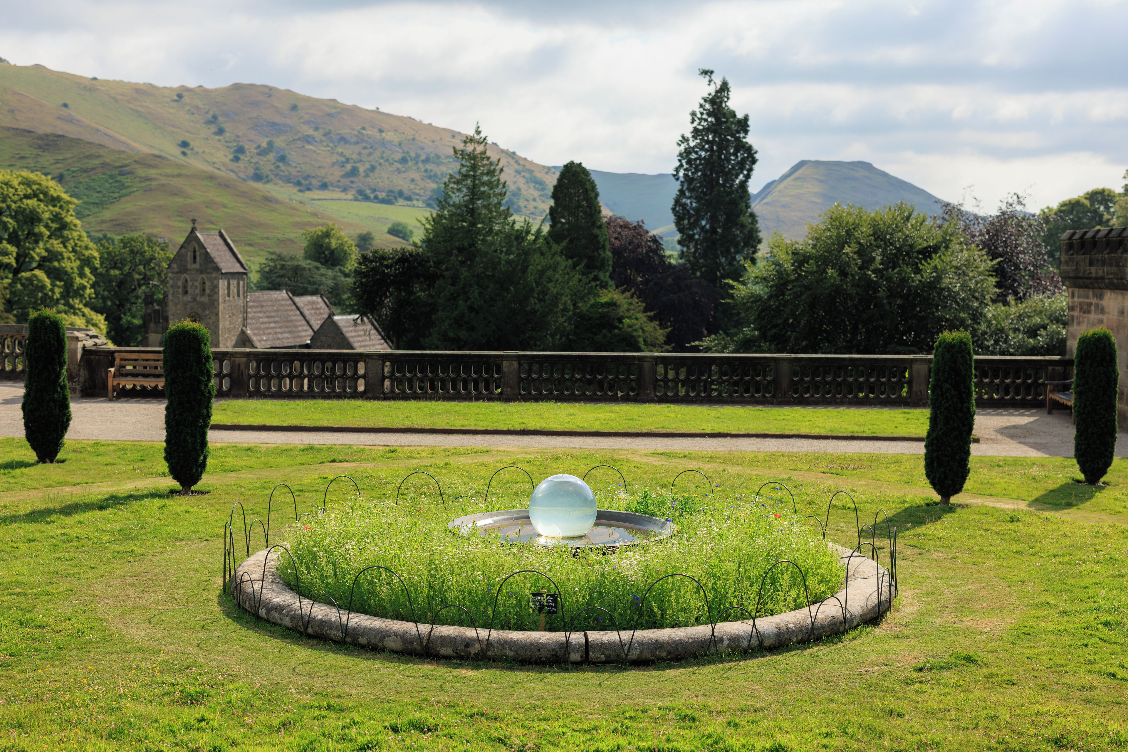 A garden with a spherical water feature, a stone wall, and a distant view of a church and hills.