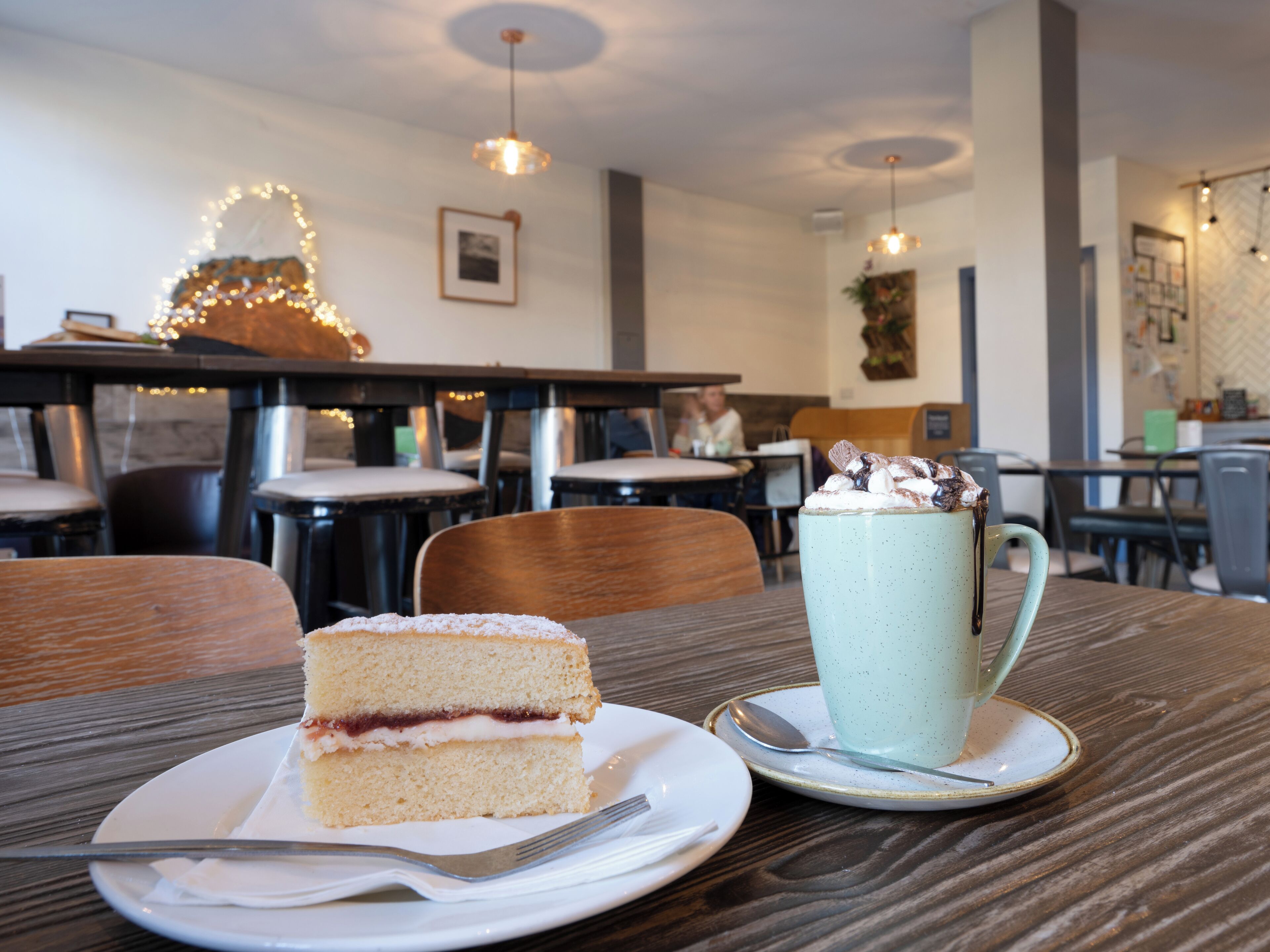 A slice of cake on a plate with a mug of hot chocolate on a wooden table in a cafe setting.