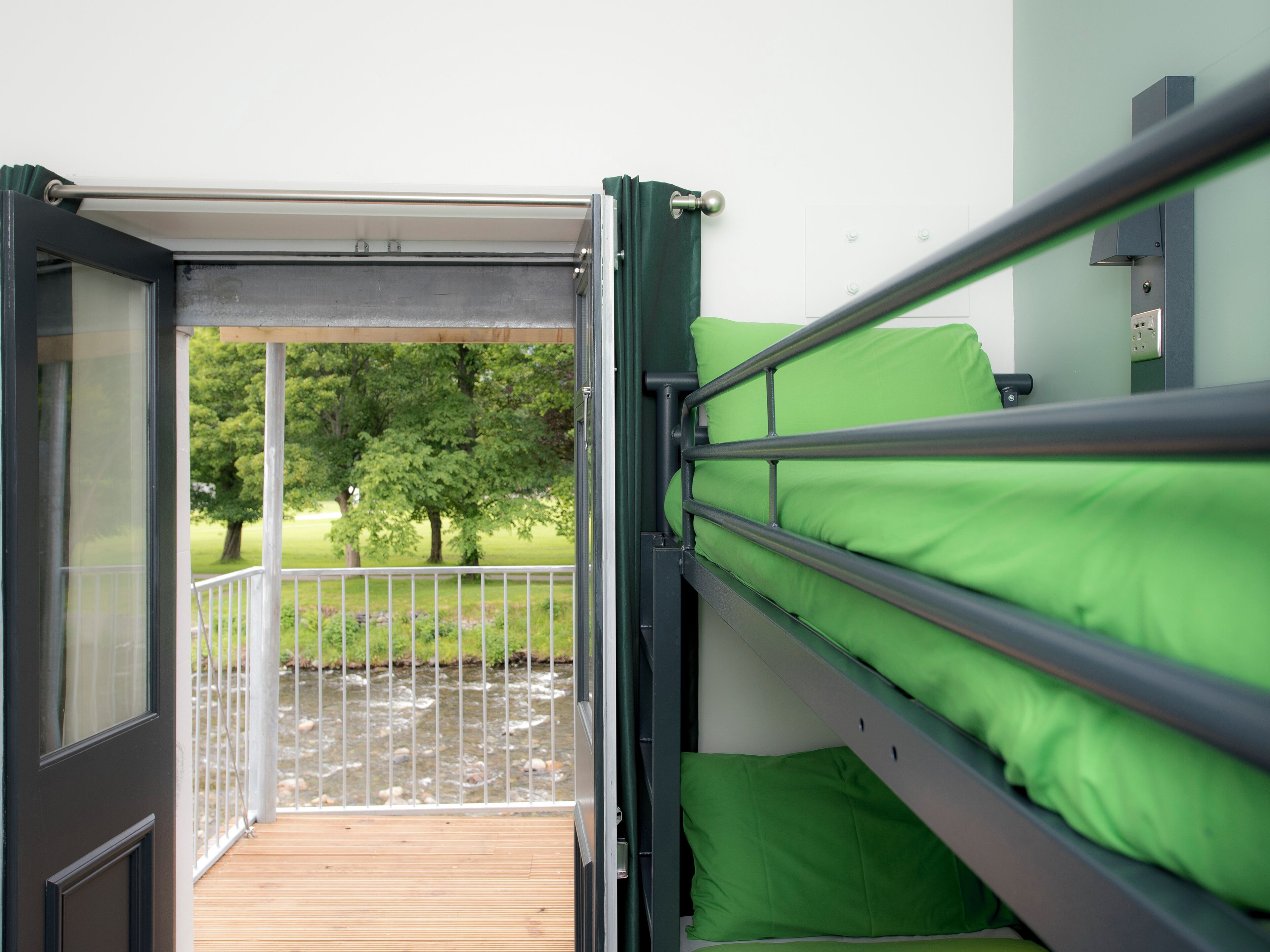 Una cama litera con ropa de cama verde, una terraza de madera y vistas a un río y árboles.