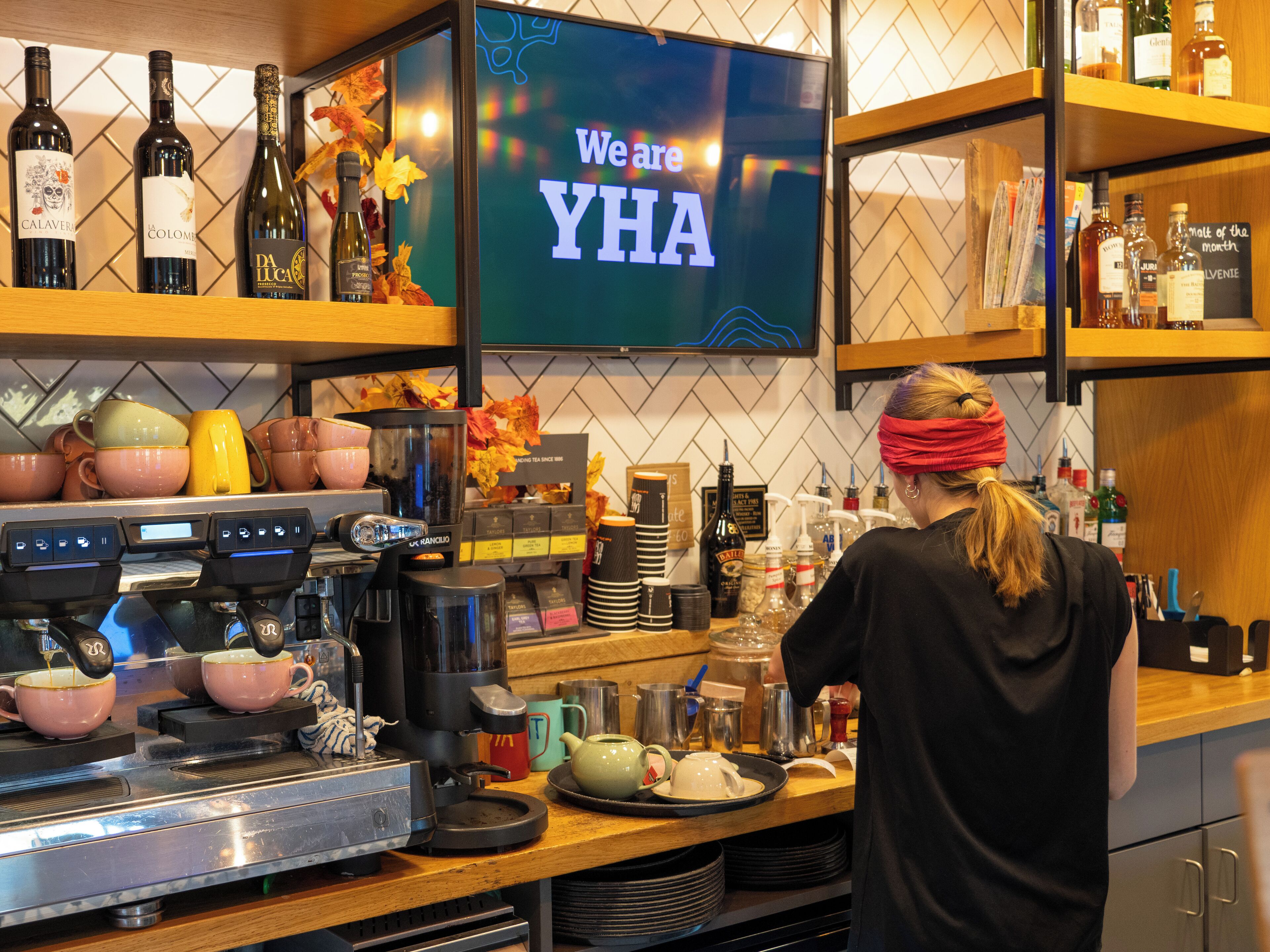 Un espace bar équipé d’une machine à café, d’étagères remplies de bouteilles et d’une enseigne indiquant : « Nous sommes YHA ».