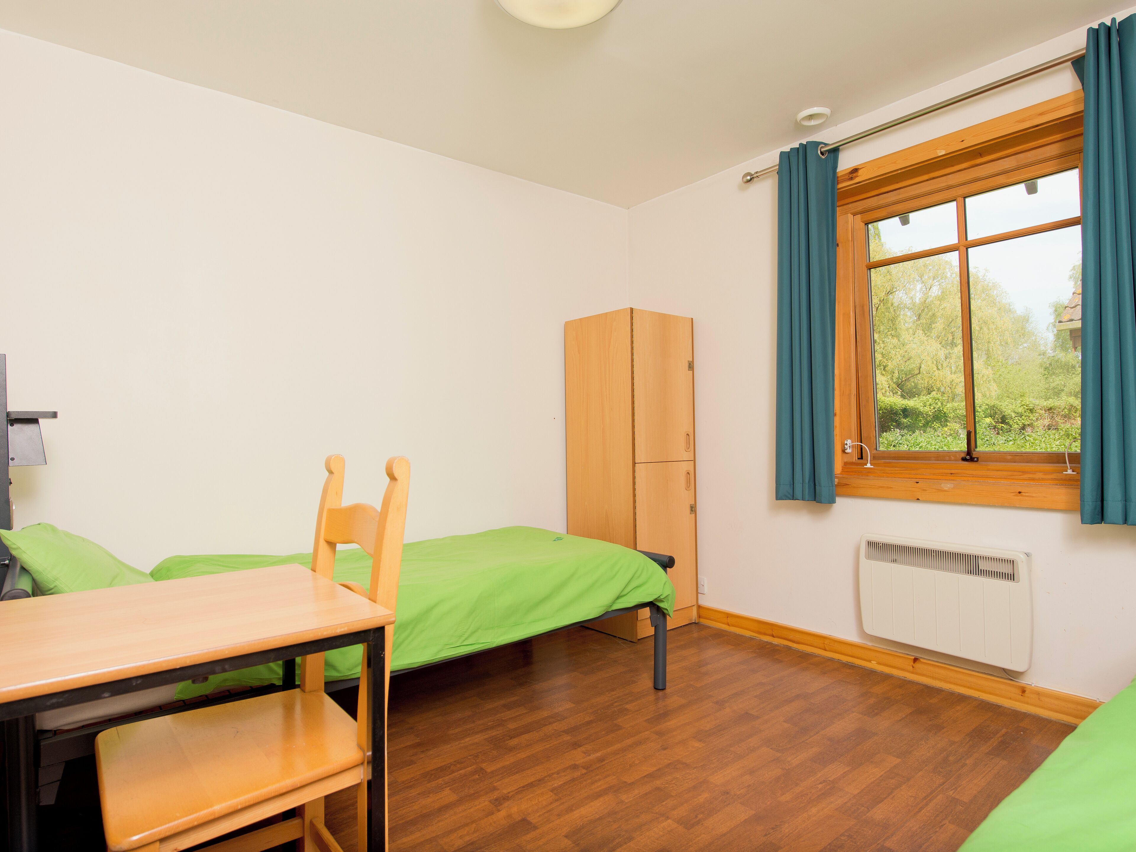 A small, tidy room with a wooden desk, two single beds, a wardrobe, and a window with curtains.
