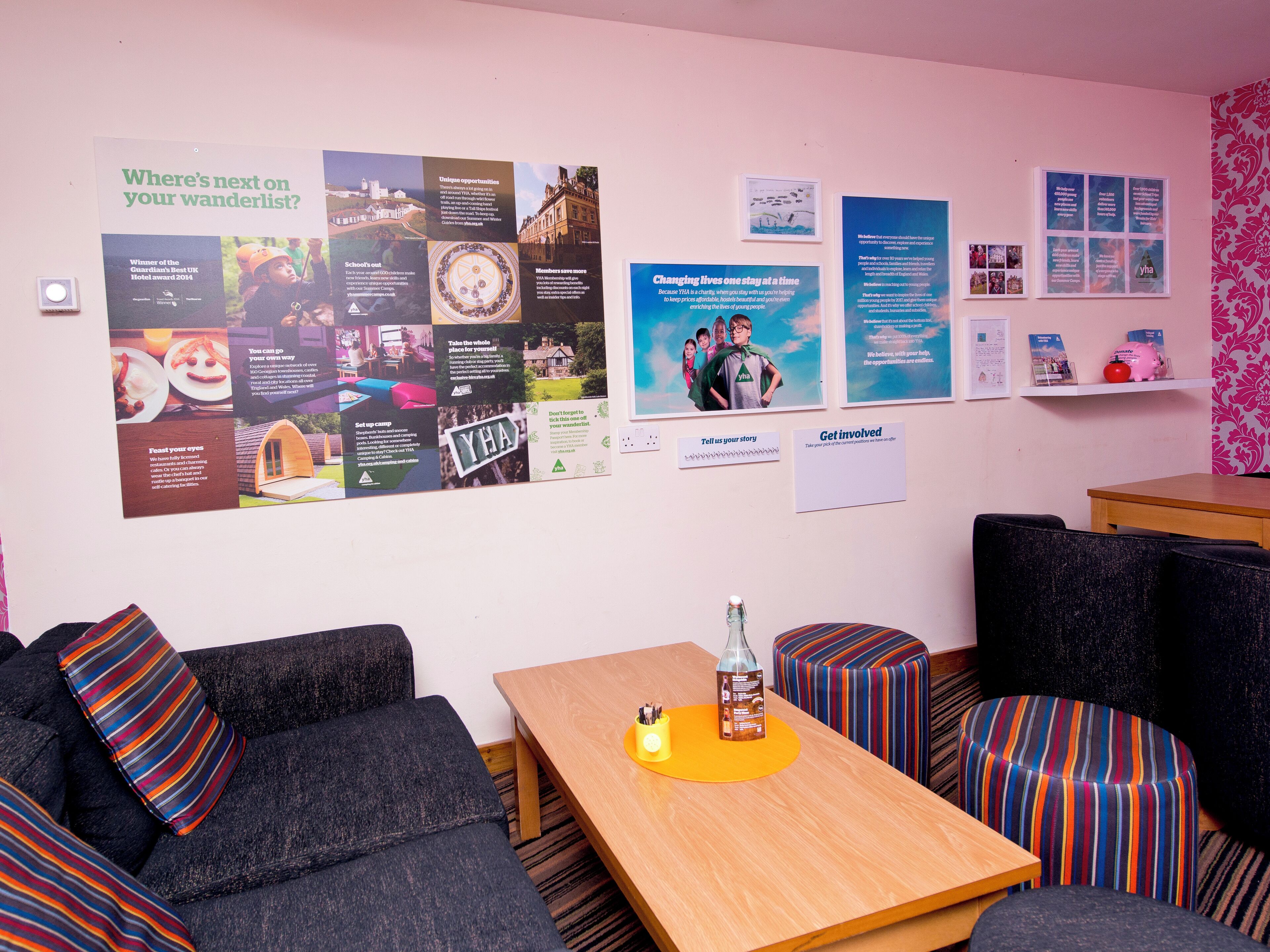 Une pièce chaleureuse avec une table en bois, des coussins à rayures et un panneau d’affichage contenant des informations sur les voyages.
