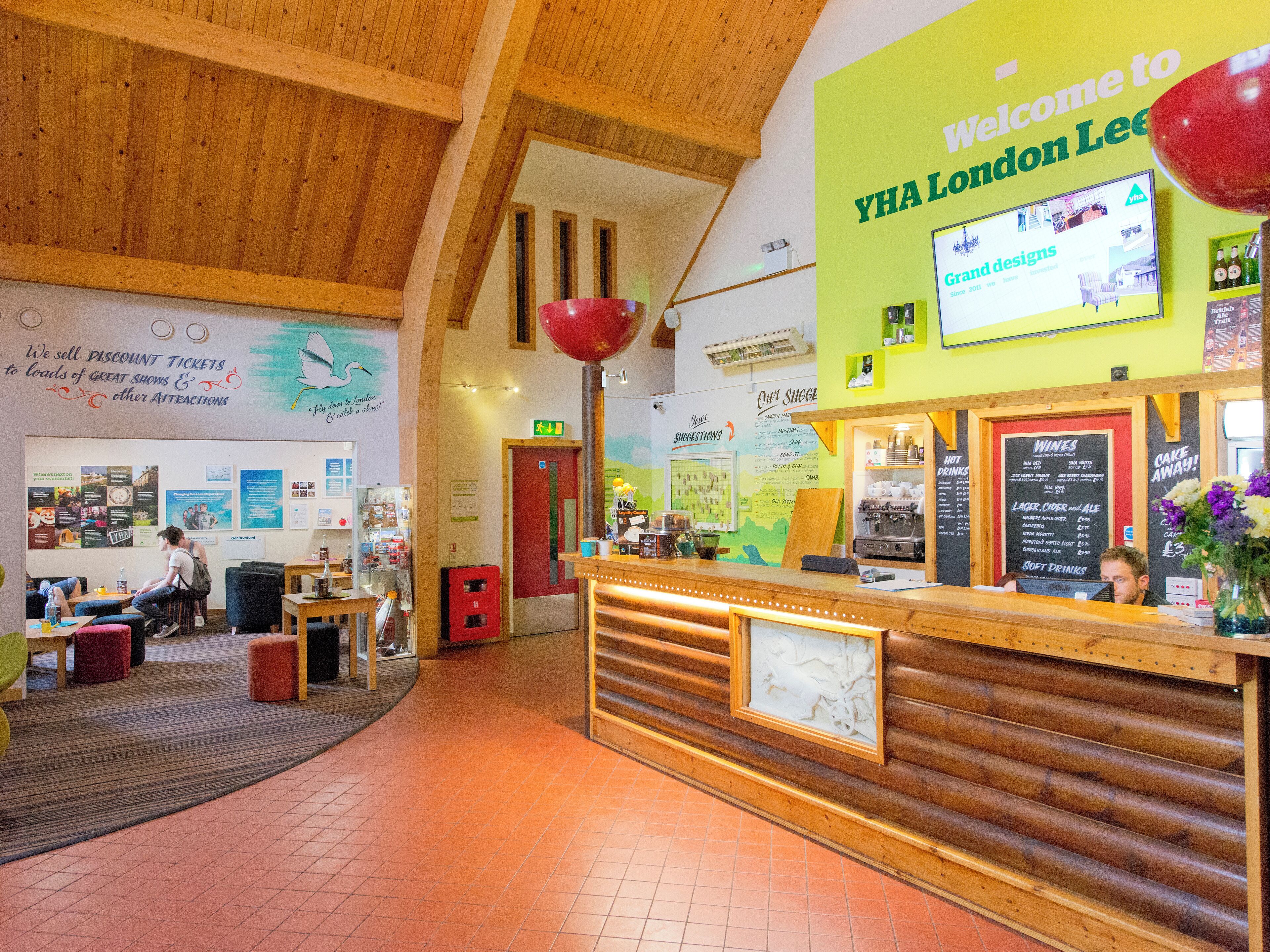 Un espace d’accueil avec un comptoir en bois, un écran mural affichant des informations et un panneau accueillant les visiteurs à la auberge de jeunesse YHA London Lee.