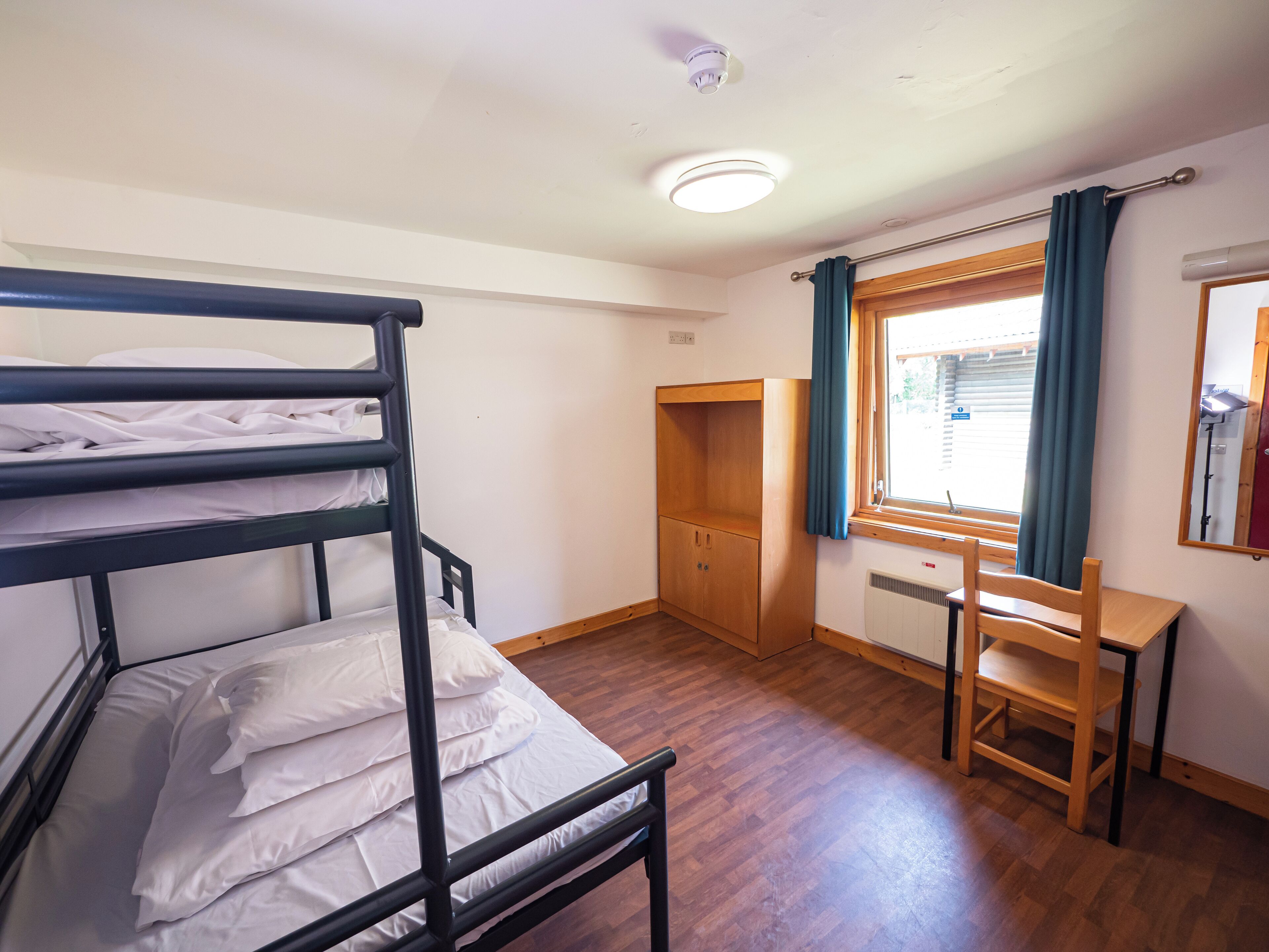 A bunk bed with white bedding, a wooden desk, a chair, a wardrobe, and a window with curtains.