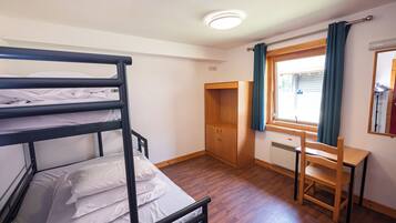 A bunk bed with white bedding, a wooden desk, a chair, a wardrobe, and a window with curtains.