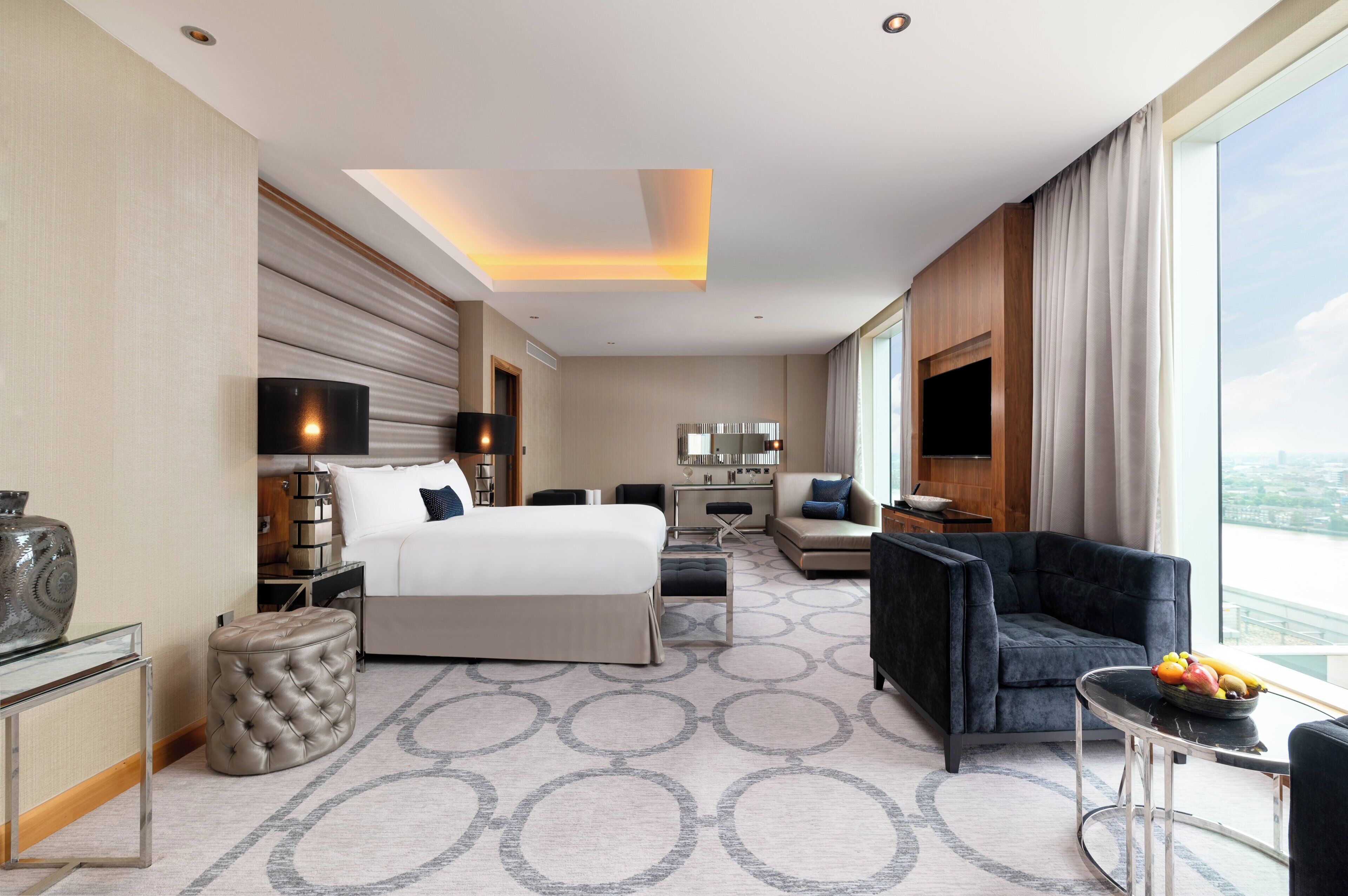A modern hotel room with a large bed, a sofa, a TV, and a dining area with a fruit bowl.