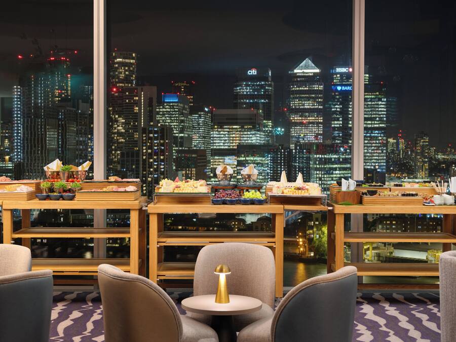 Habitación de hotel en un edificio alto con vista al horizonte urbano, una mesa de comedor con comida y muebles modernos.