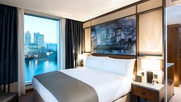 A hotel room with a large bed, a view of a cityscape, a painting on the wall, and a nightstand with a lamp.