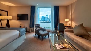 A hotel room with a bed, two armchairs, a sofa, a glass coffee table, a TV, and a view of a city skyline.