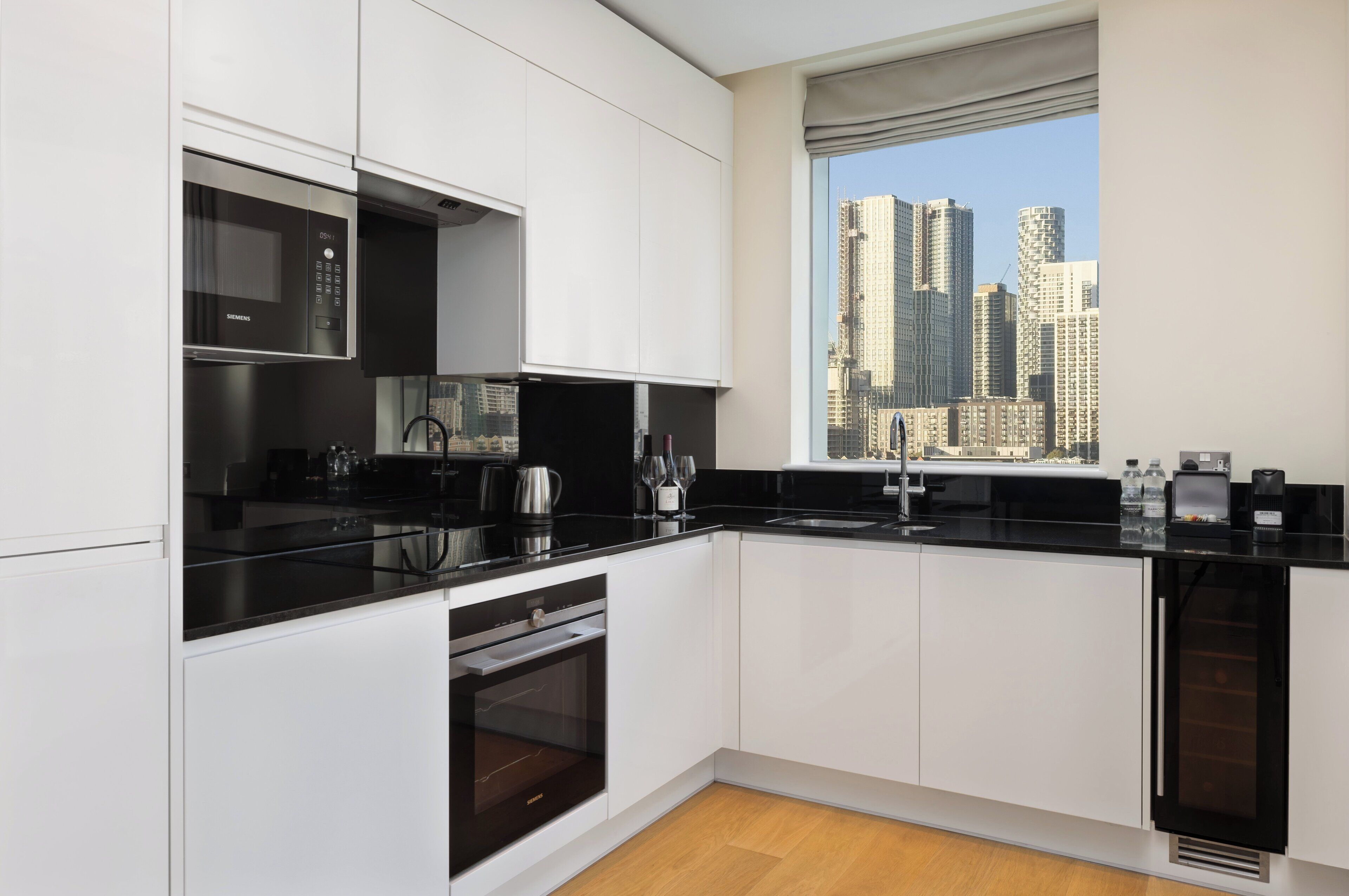 A modern kitchen with white cabinetry, black countertops, and a built-in oven. A window offers a view of tall buildings.