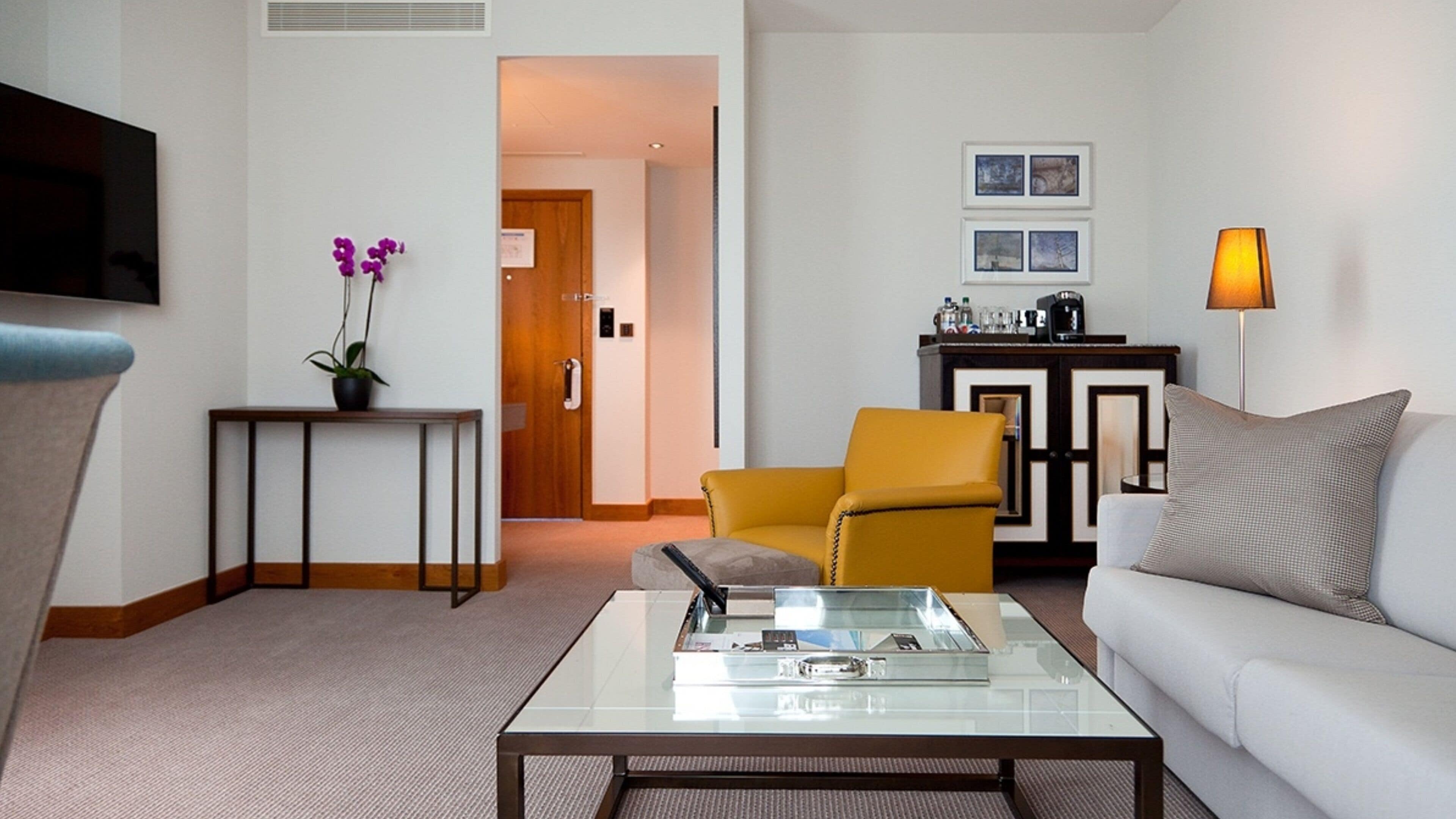 Een moderne woonkamer met een glazen salontafel, een bank, een gele fauteuil, een kleine tafel met een bloempot en een flatscreen televisie aan de muur.
