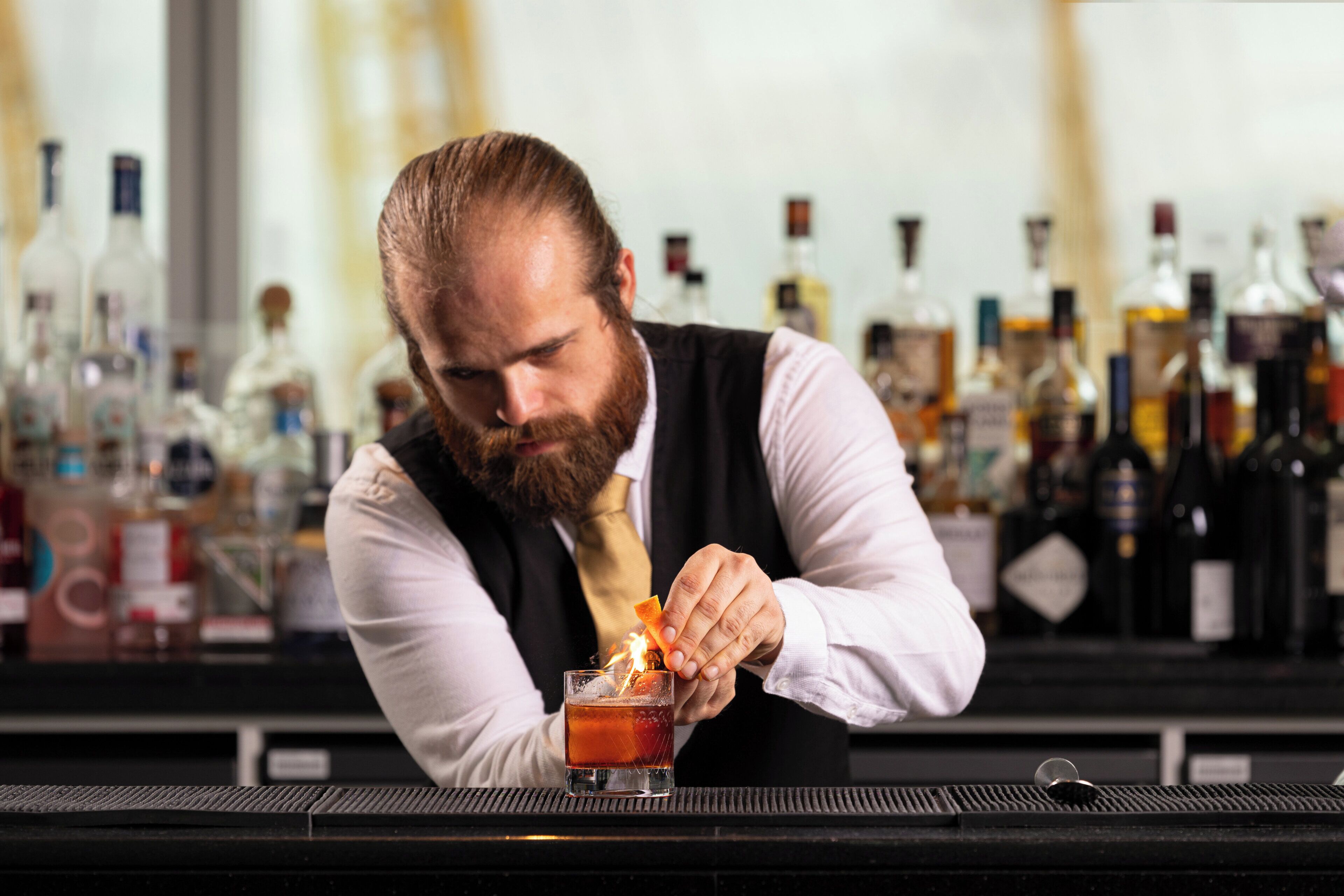 A bartender preparing a drink at a bar with various bottles in the background.
