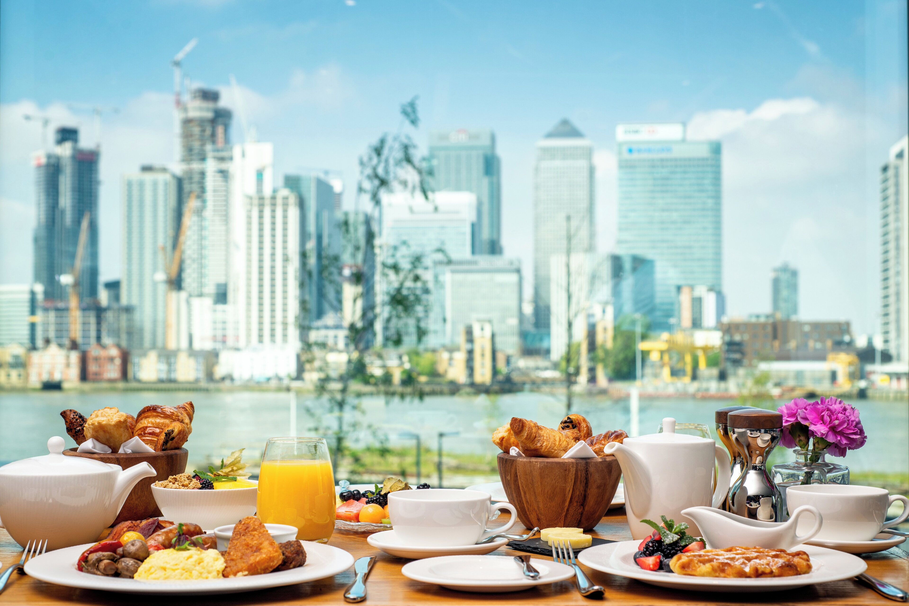 A breakfast table with croissants, fruit, and a glass of orange juice, set against a city skyline view.