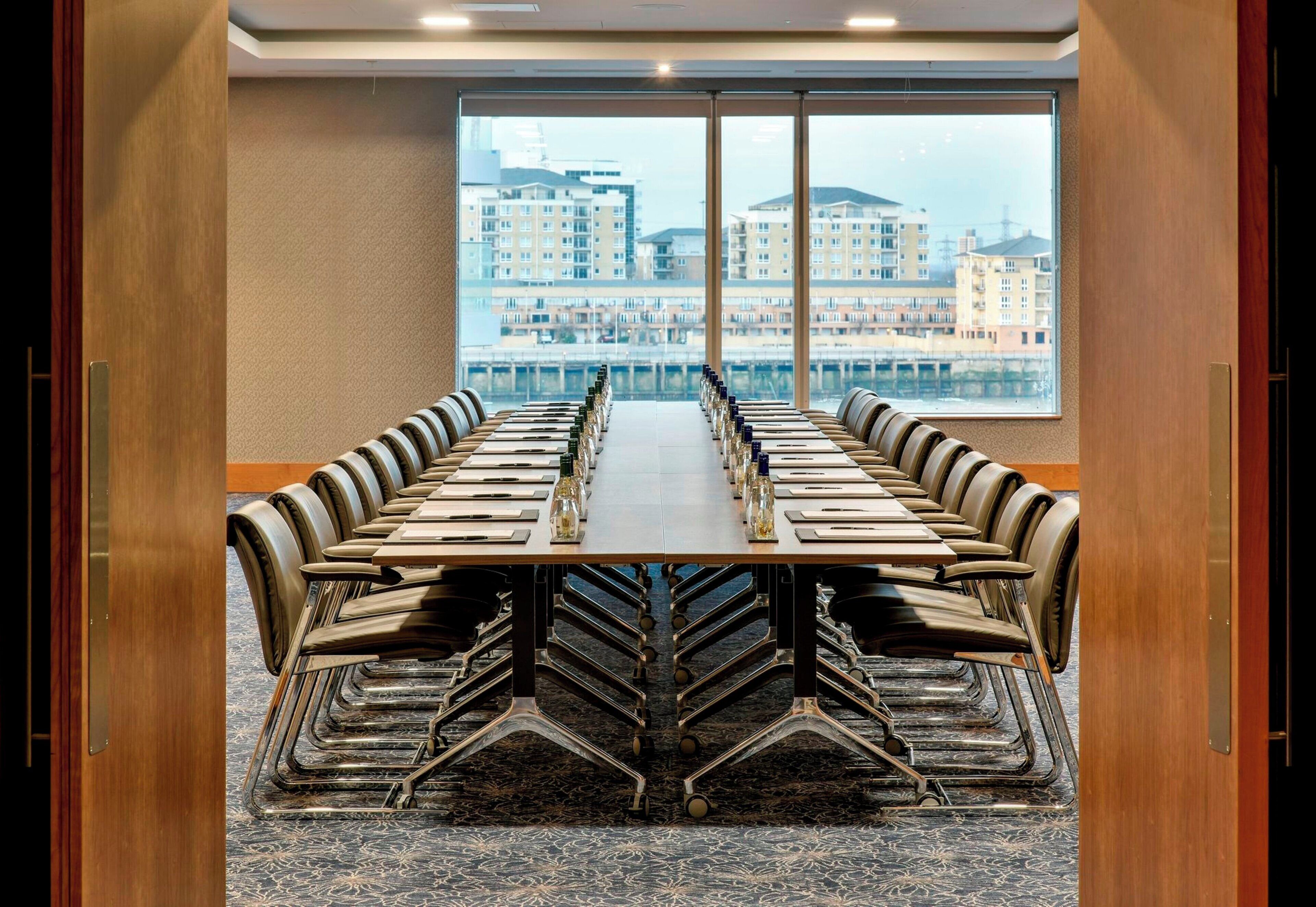 Sala de conferências com uma mesa comprida e cadeiras, amplas janelas com vista para edifícios e piso coberto de carpete.