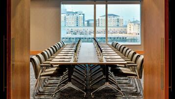 Sala de conferências com uma mesa comprida e cadeiras, amplas janelas com vista para edifícios e piso coberto de carpete.