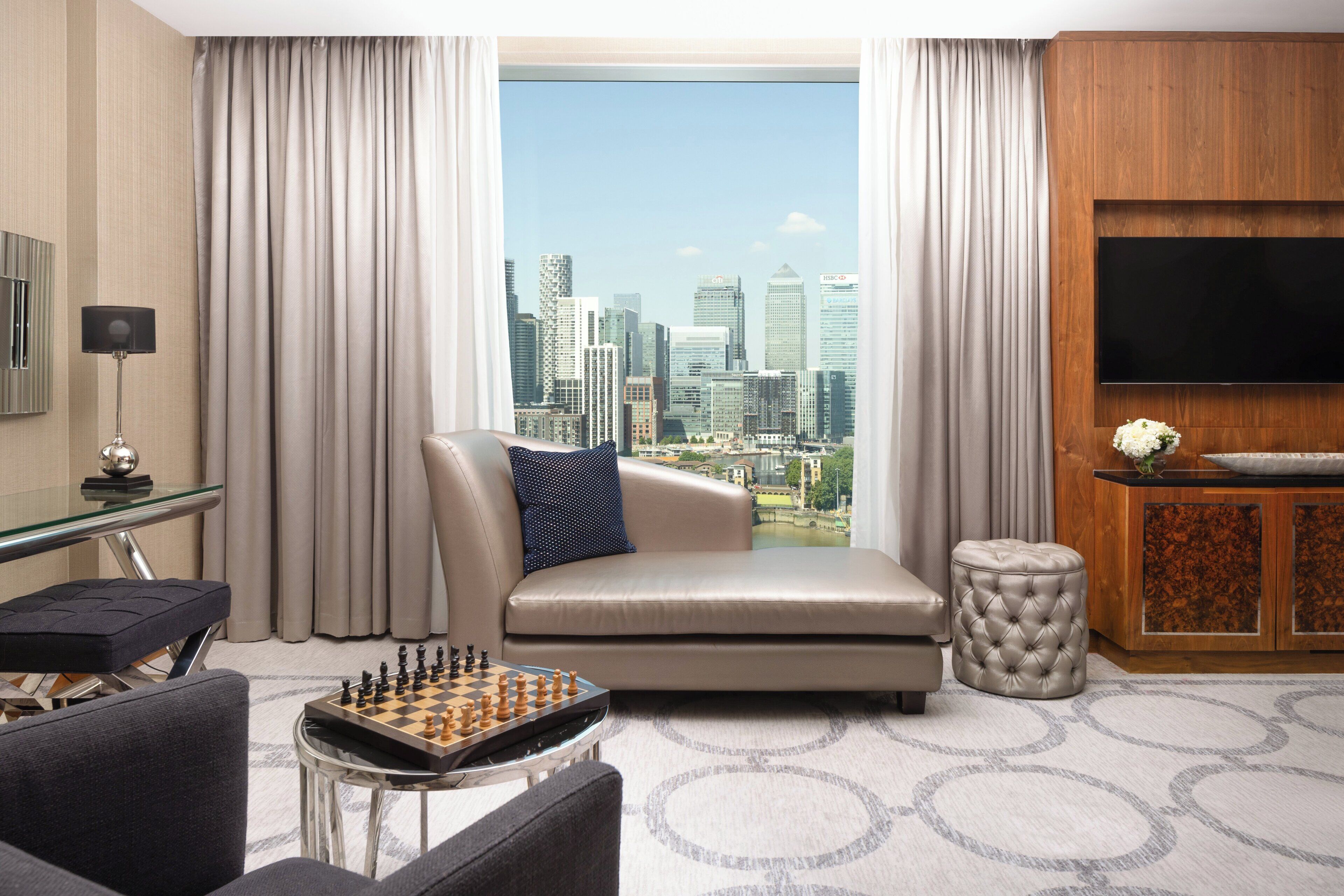 Une chambre d’hôtel moderne avec vue sur la ville, comprenant un canapé, un téléviseur, une table d’échecs et un petit pouf.