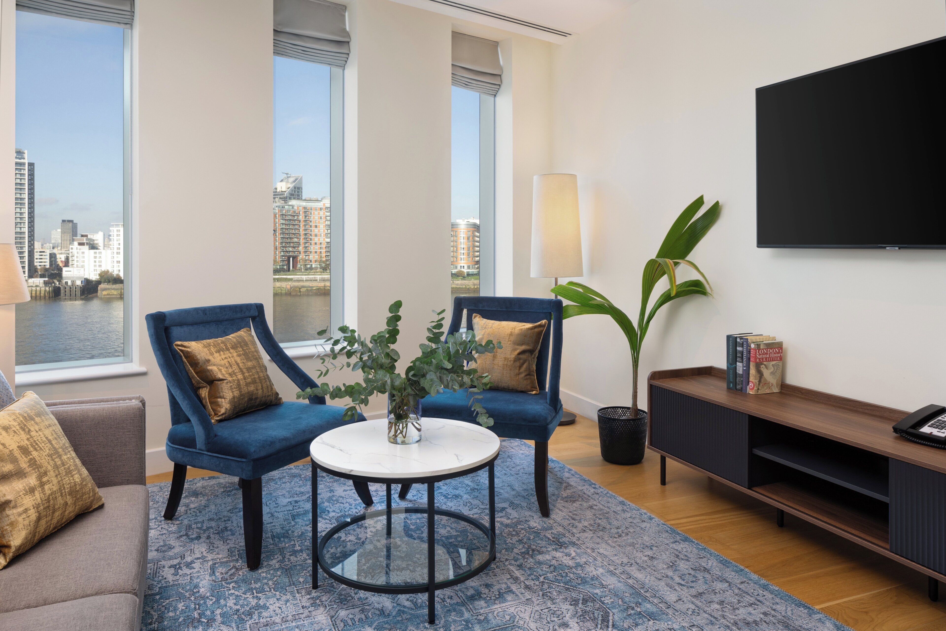 A modern living room with two blue armchairs, a white coffee table, a bookshelf, and a large window overlooking a cityscape.