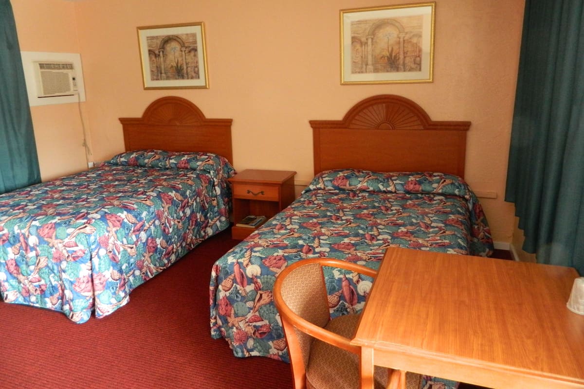 A hotel room with two beds, a wooden nightstand, a chair, a table, and two framed pictures on the wall.